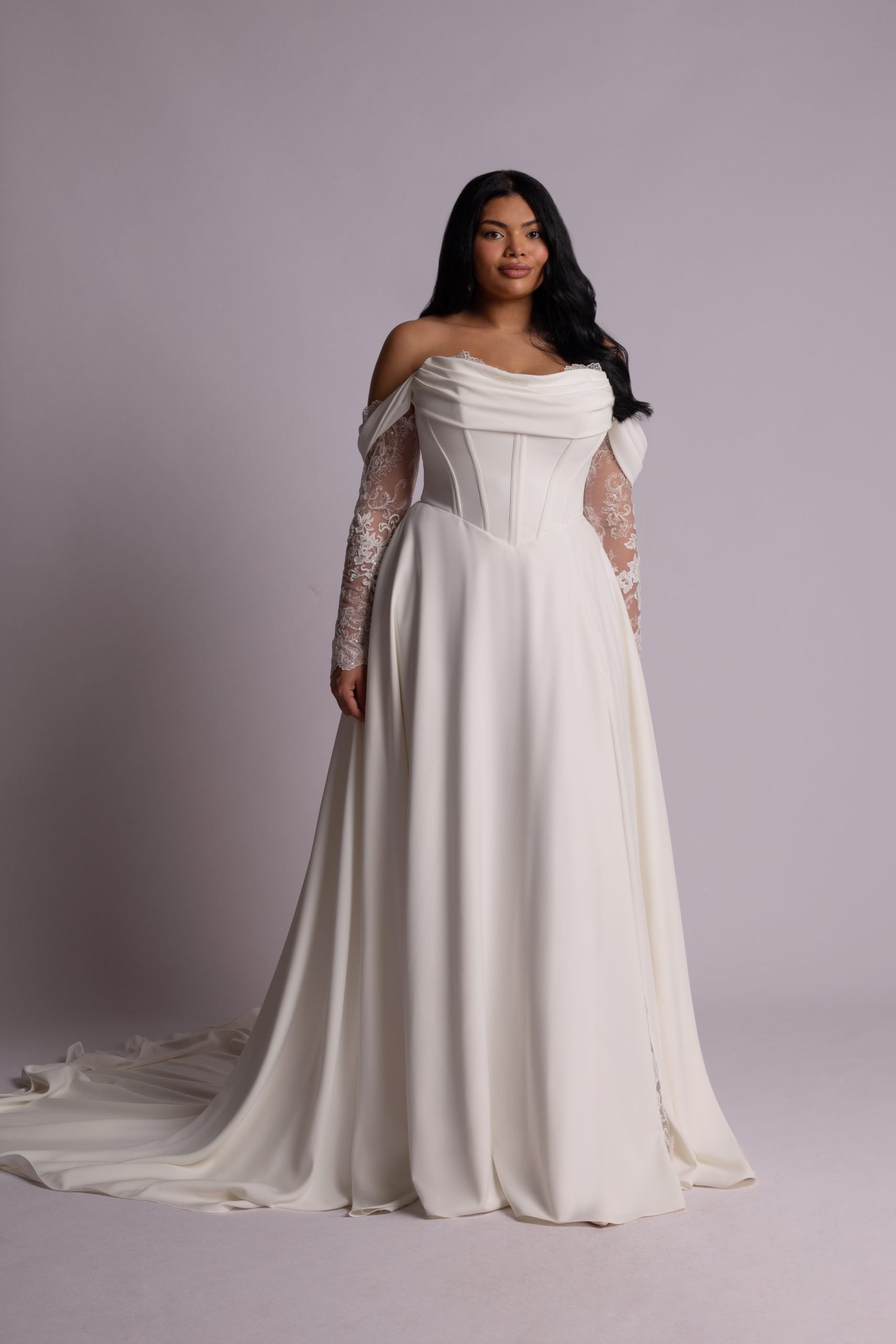 A woman in a white wedding dress with lace sleeves and off-the-shoulder design, standing against a plain light gray background.