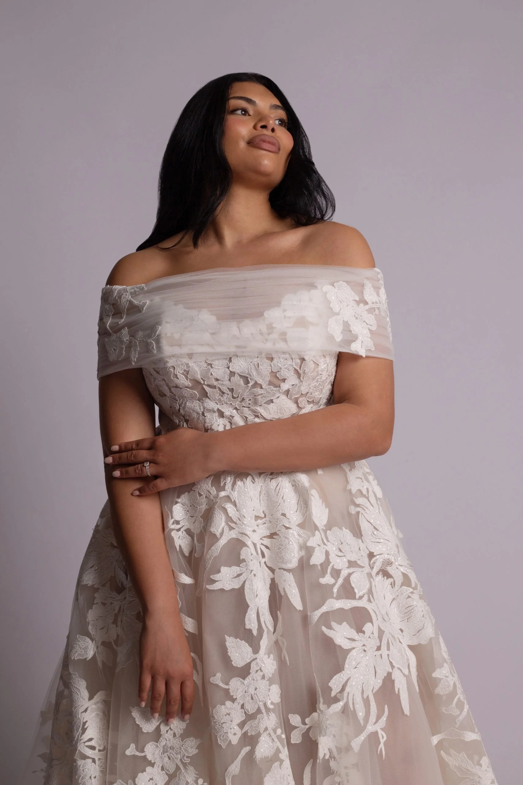 A woman in a strapless, off-the-shoulder wedding dress with lace and floral embroidery, standing against a plain light gray background.