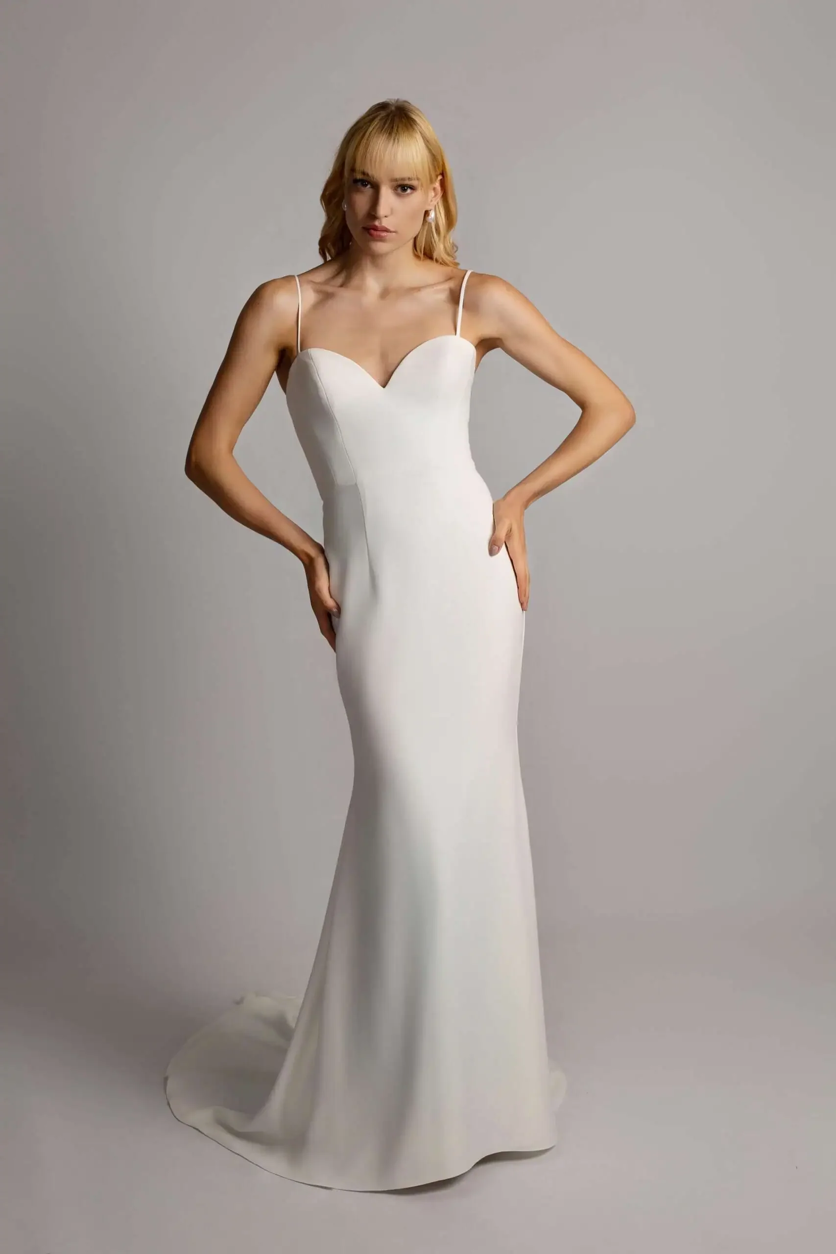Woman in a fitted white wedding dress with spaghetti straps posing against a plain gray background.