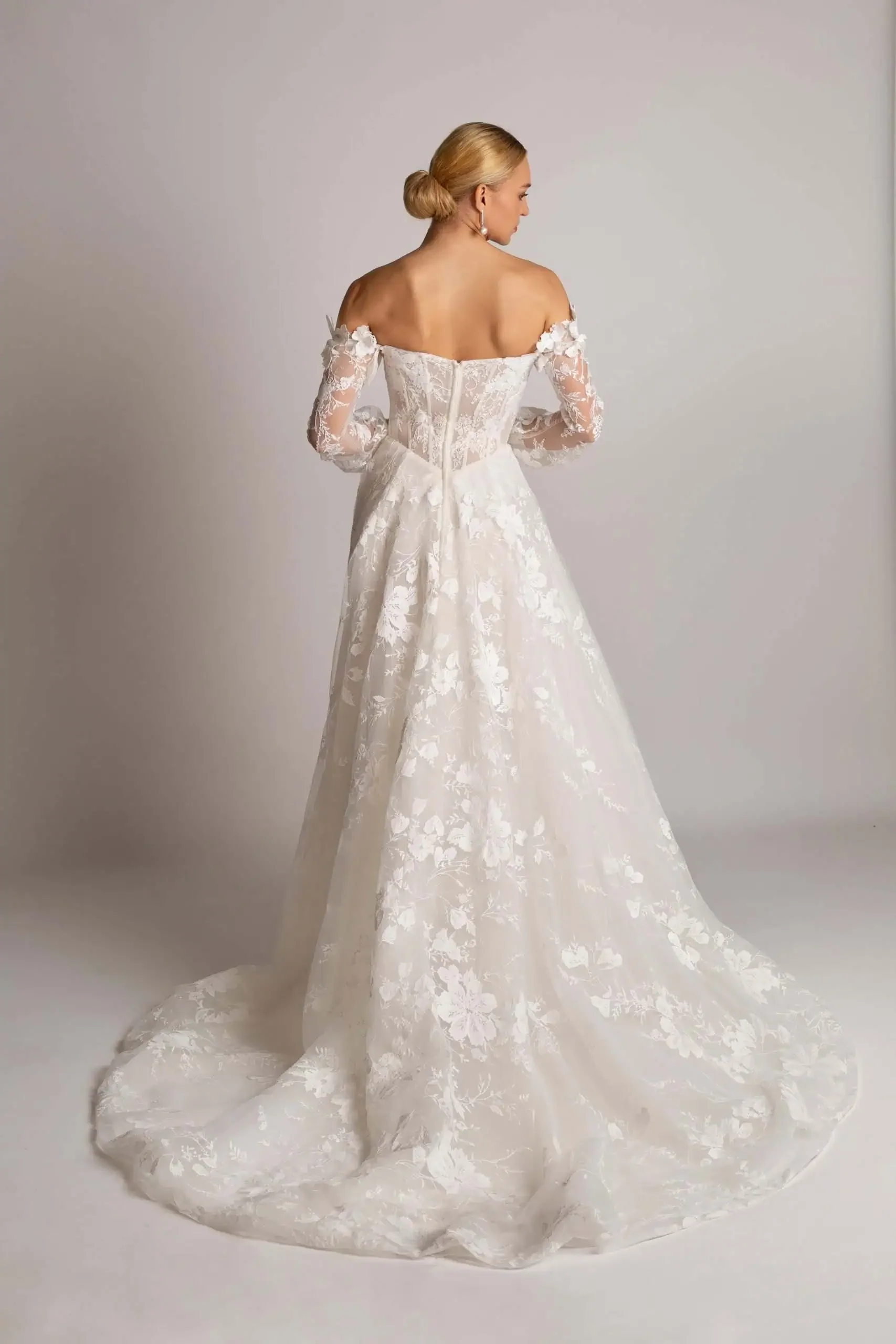 A woman in a white, off-the-shoulder, lace wedding gown with a long train, standing with her back to the camera against a plain neutral background.