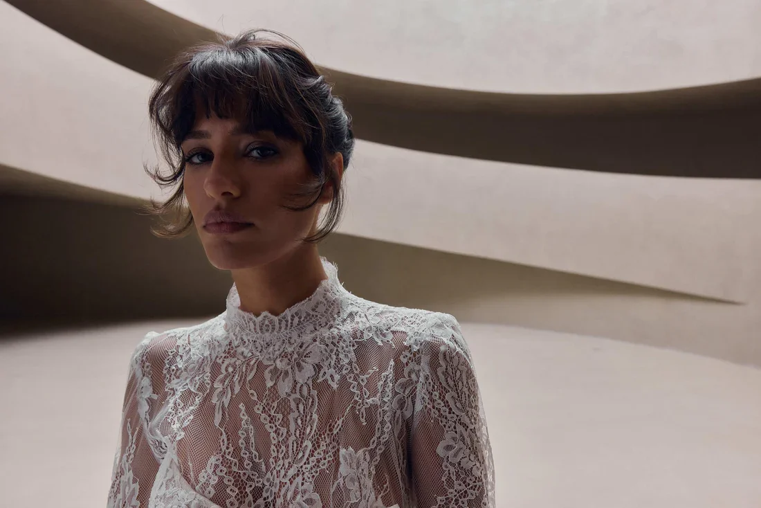 A woman with short, wavy dark hair and bangs wearing a white lace high-neck dress in front of a modern, abstract concrete architectural background.