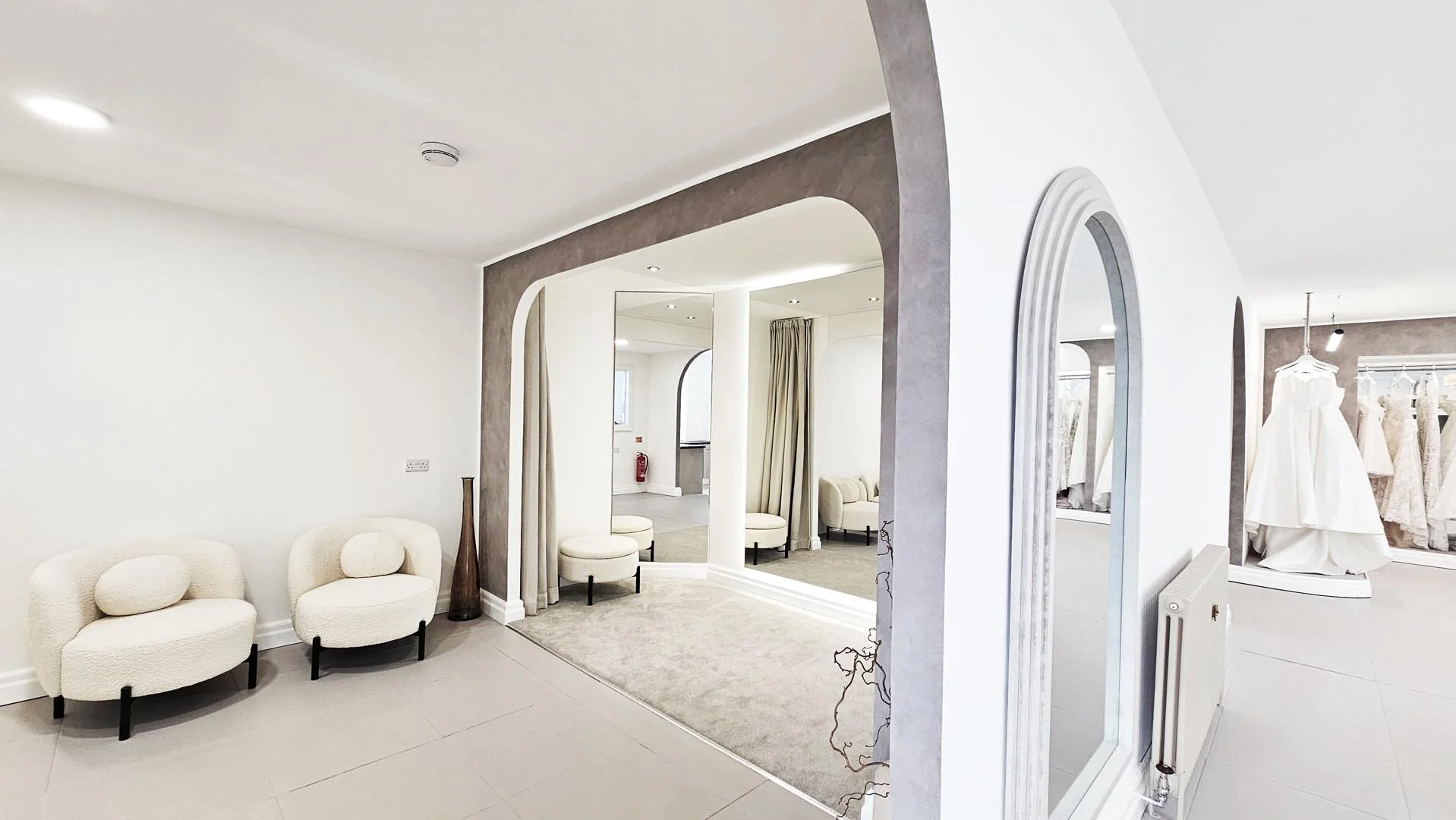 Interior of a boutique store with white walls and furniture, including two rounded white chairs and a tall brown vase, displaying wedding dresses on mannequins and racks.