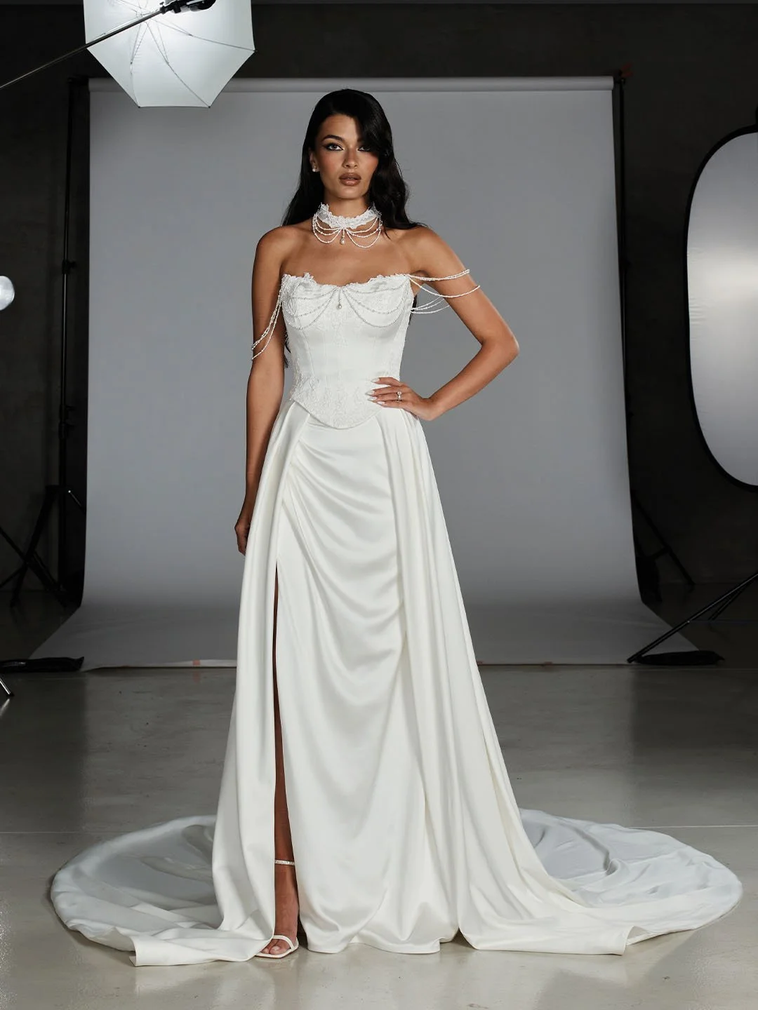 A woman in a wedding dress posing in a photography studio with a gray backdrop and studio lighting.