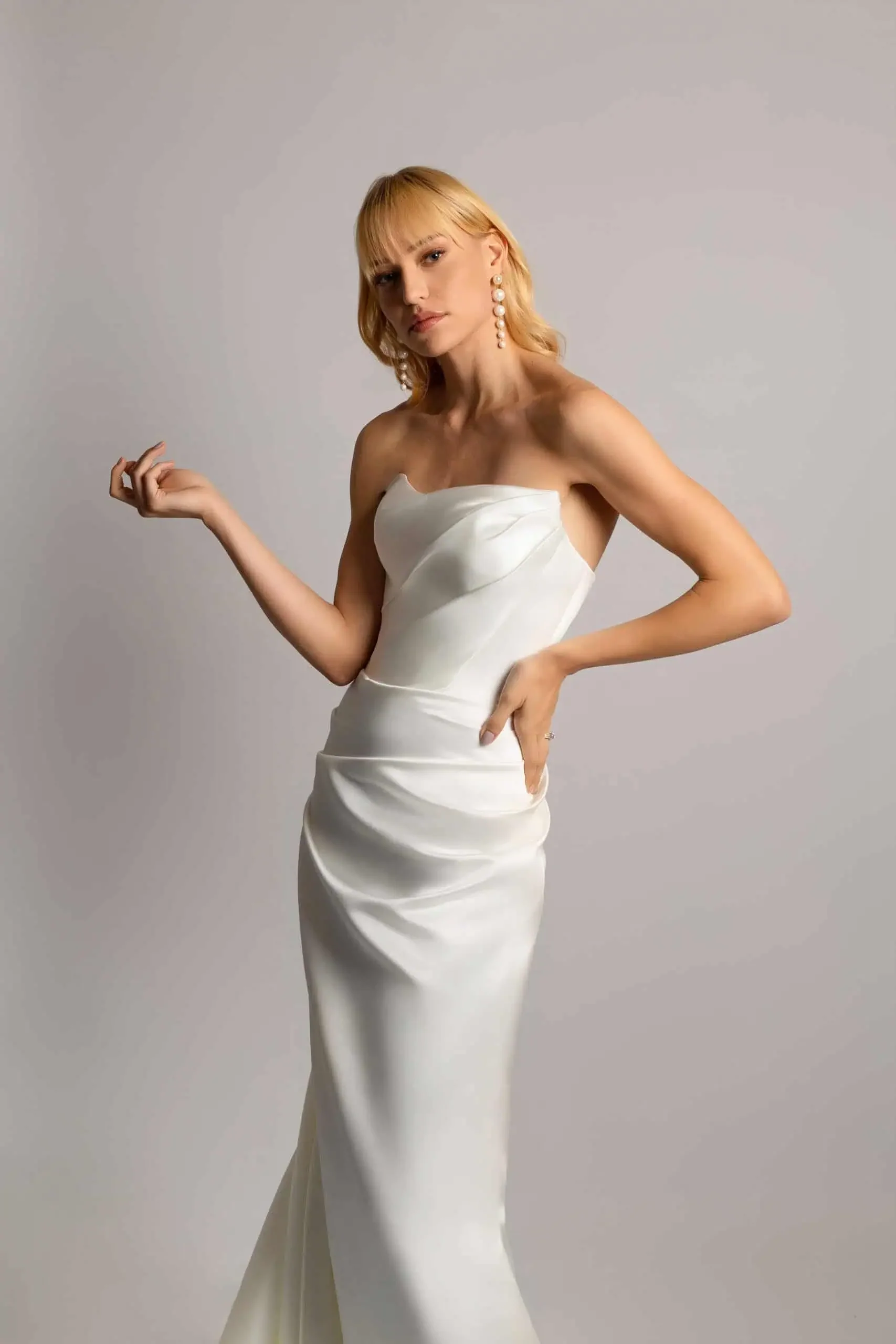 A woman with blond hair, wearing a strapless white gown with draped fabric, standing against a plain gray background.