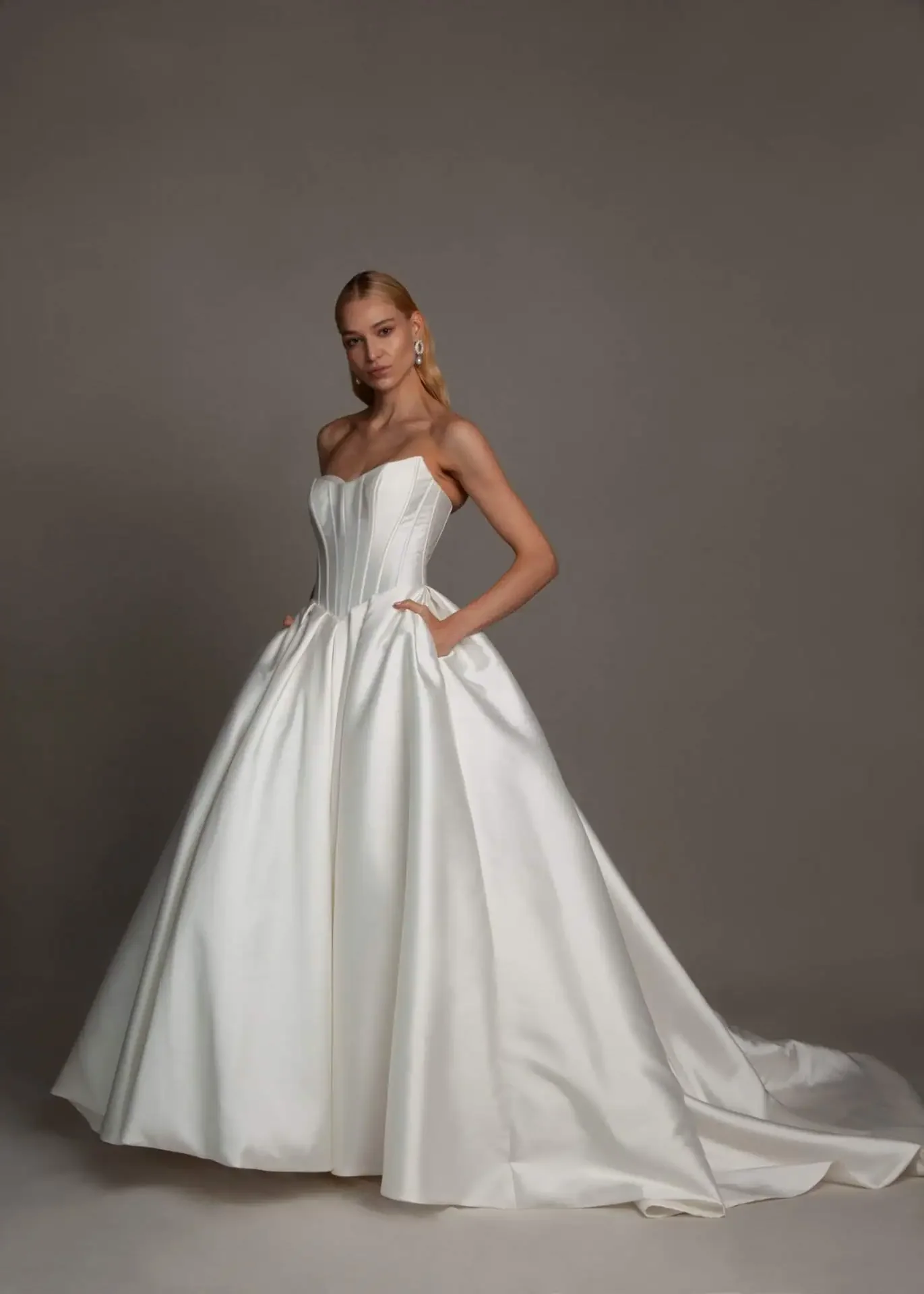 A woman wearing a strapless white wedding gown with a full skirt and a slight train, standing against a plain gray background.
