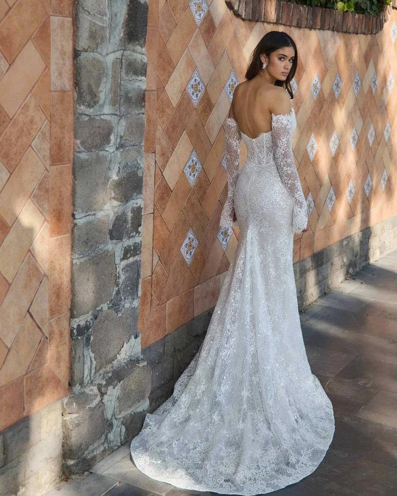 A woman in an elegant white lace wedding gown with long sleeves, standing with her back to the camera, against a tiled wall with stone accents.