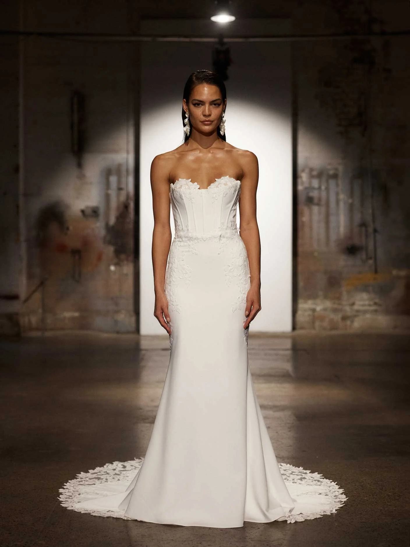 A woman in a white strapless wedding dress with lace detailing, standing on a runway with a spotlight shining behind her in an industrial-style setting.
