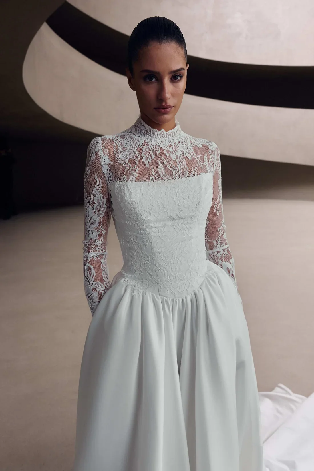A woman wearing a white lace wedding dress with long sleeves, standing indoors in front of a modern, curved wall.