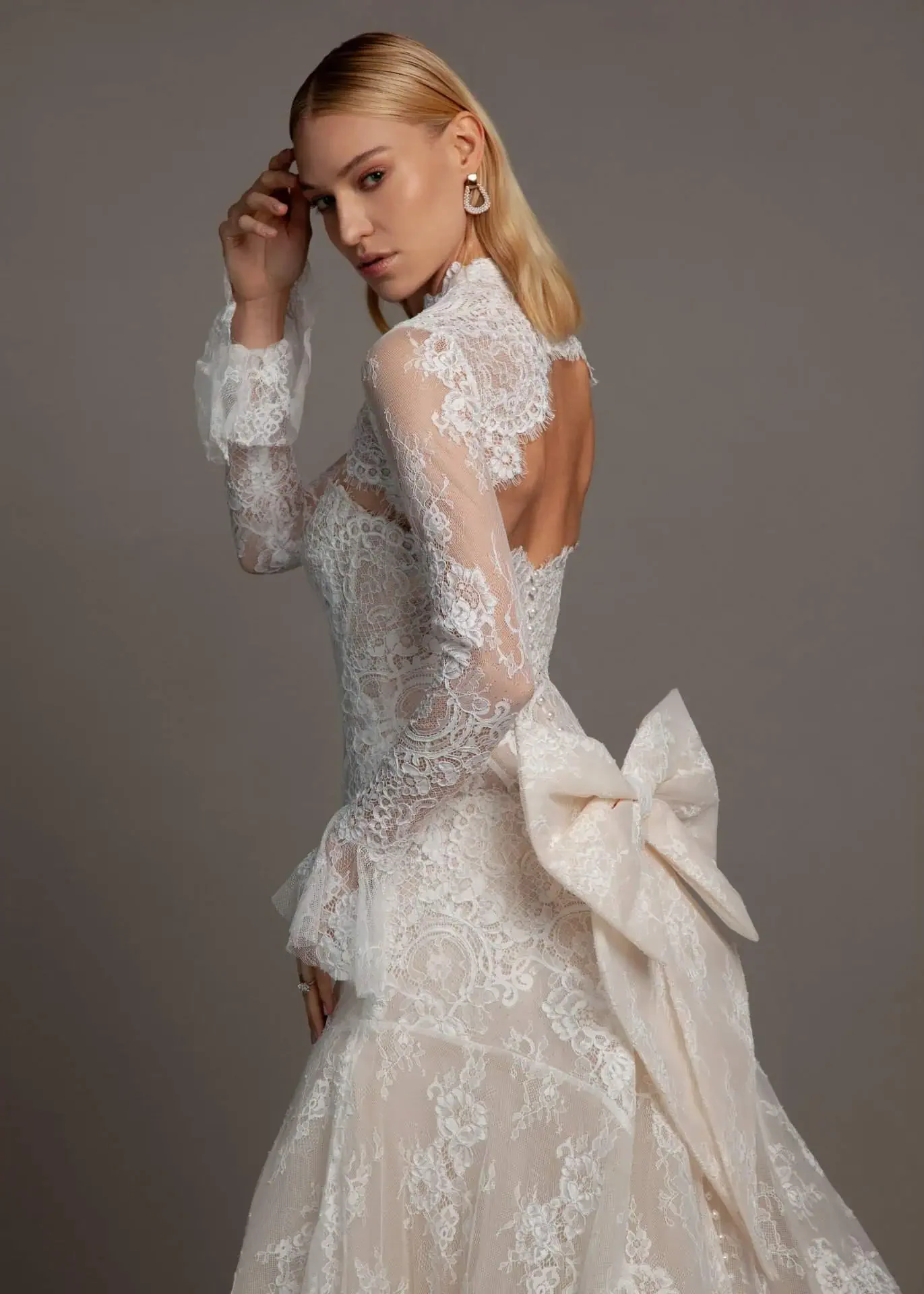 A woman with blonde hair wearing a white lace wedding dress with a large bow on the back, posing against a gray background.