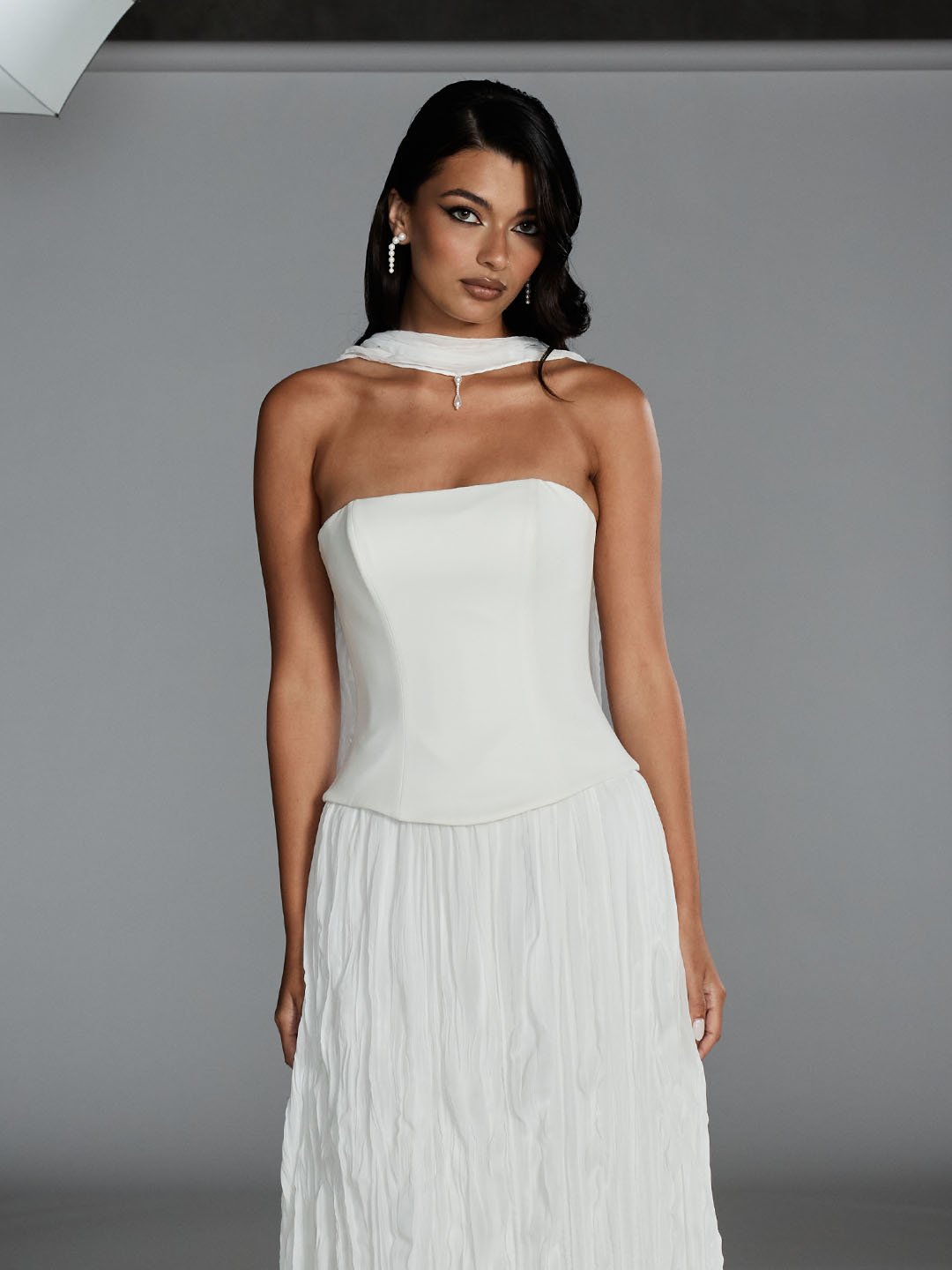 A woman in a strapless white dress with a pleated skirt, wearing pearl earrings, a choker with a pendant, and dark makeup, standing against a plain gray background.
