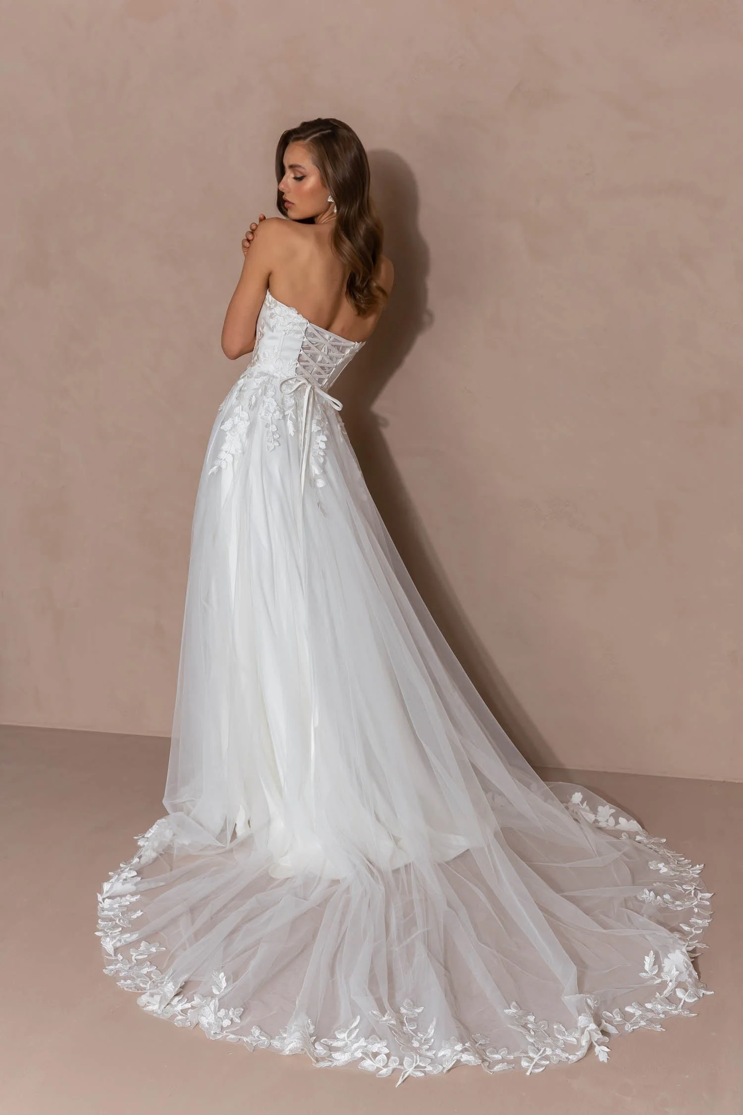 A woman in a white wedding dress with a fitted bodice, lace-up back, and a long tulle train, posing against a beige wall.