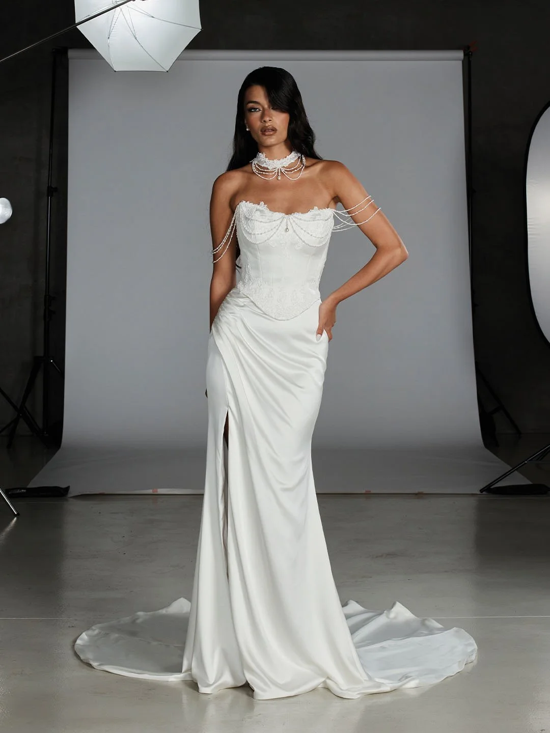 A woman in an elegant white satin wedding dress with off-the-shoulder lace and pearl jewelry posing in a photography studio with a gray backdrop.