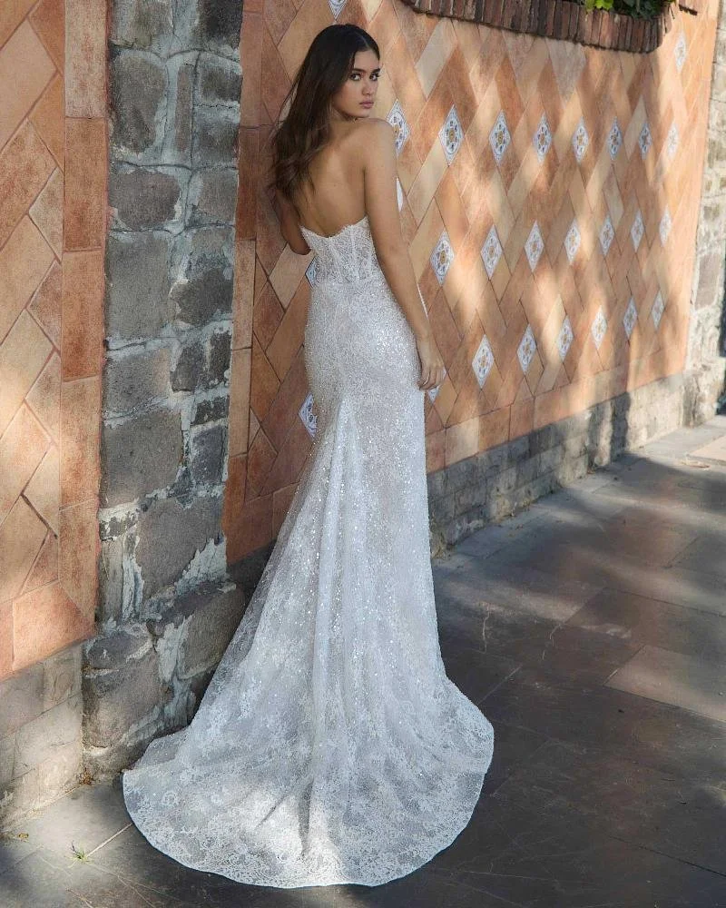 A woman in a strapless, sparkling white gown with a train, standing against a textured brick and stone wall.