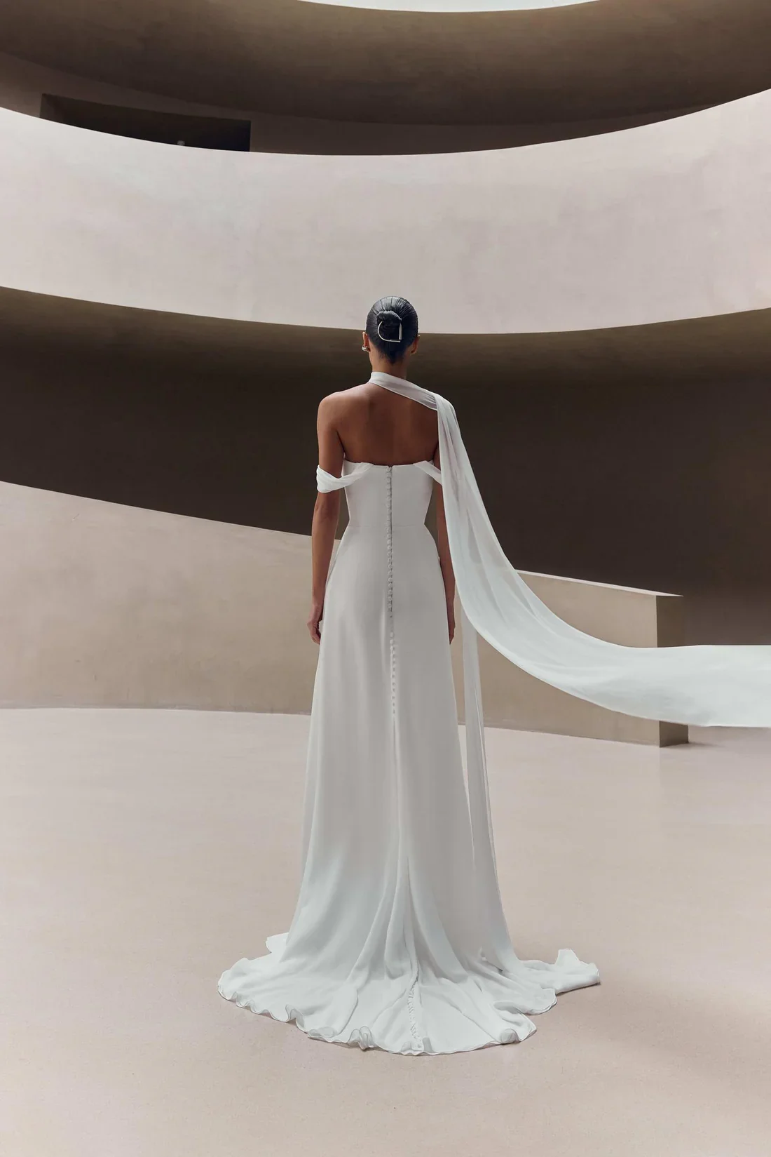 Woman in a white gown with a flowing train standing in a modern, minimalist architectural space, facing away from the camera.