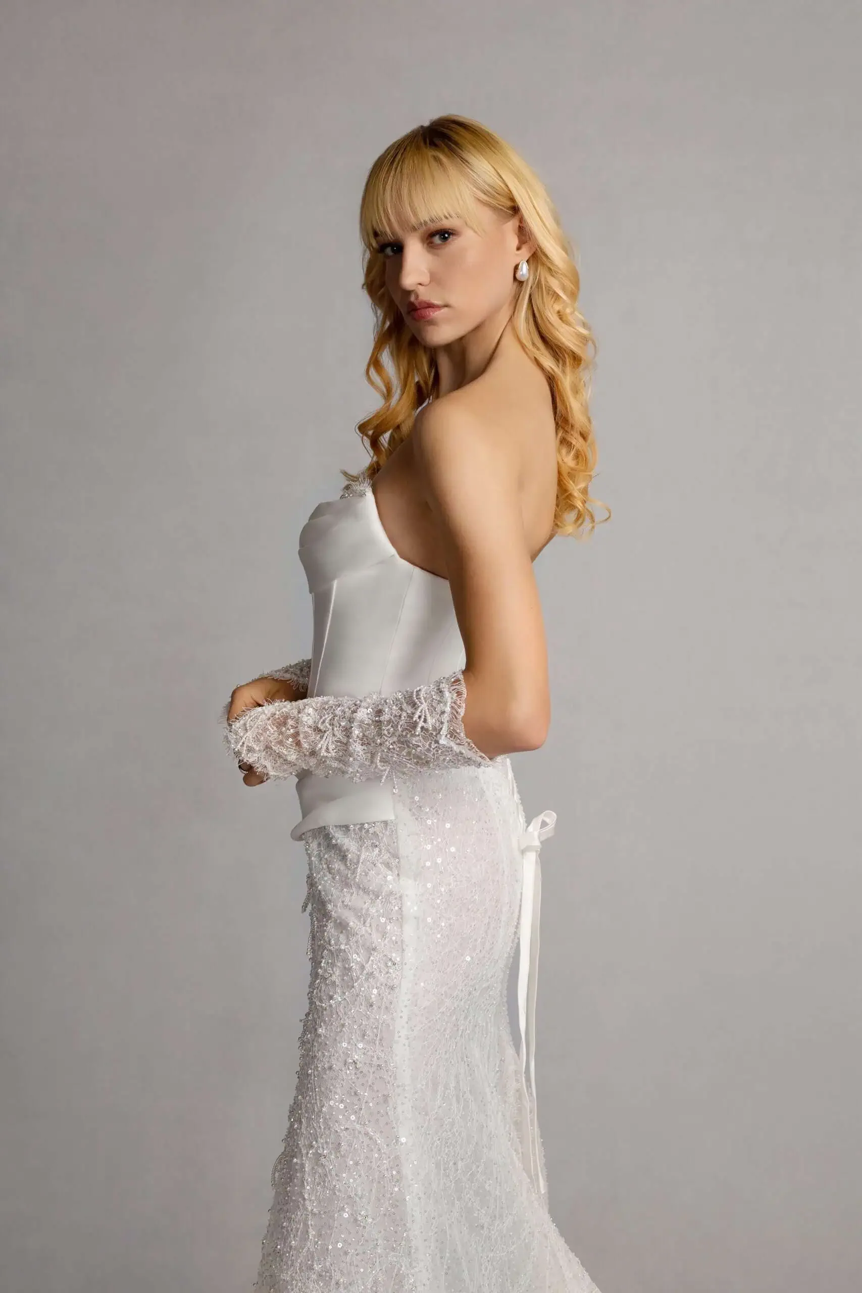 A young woman with long curly blonde hair wearing an elegant white strapless wedding dress with lace and sequin details, posing against a plain gray background.