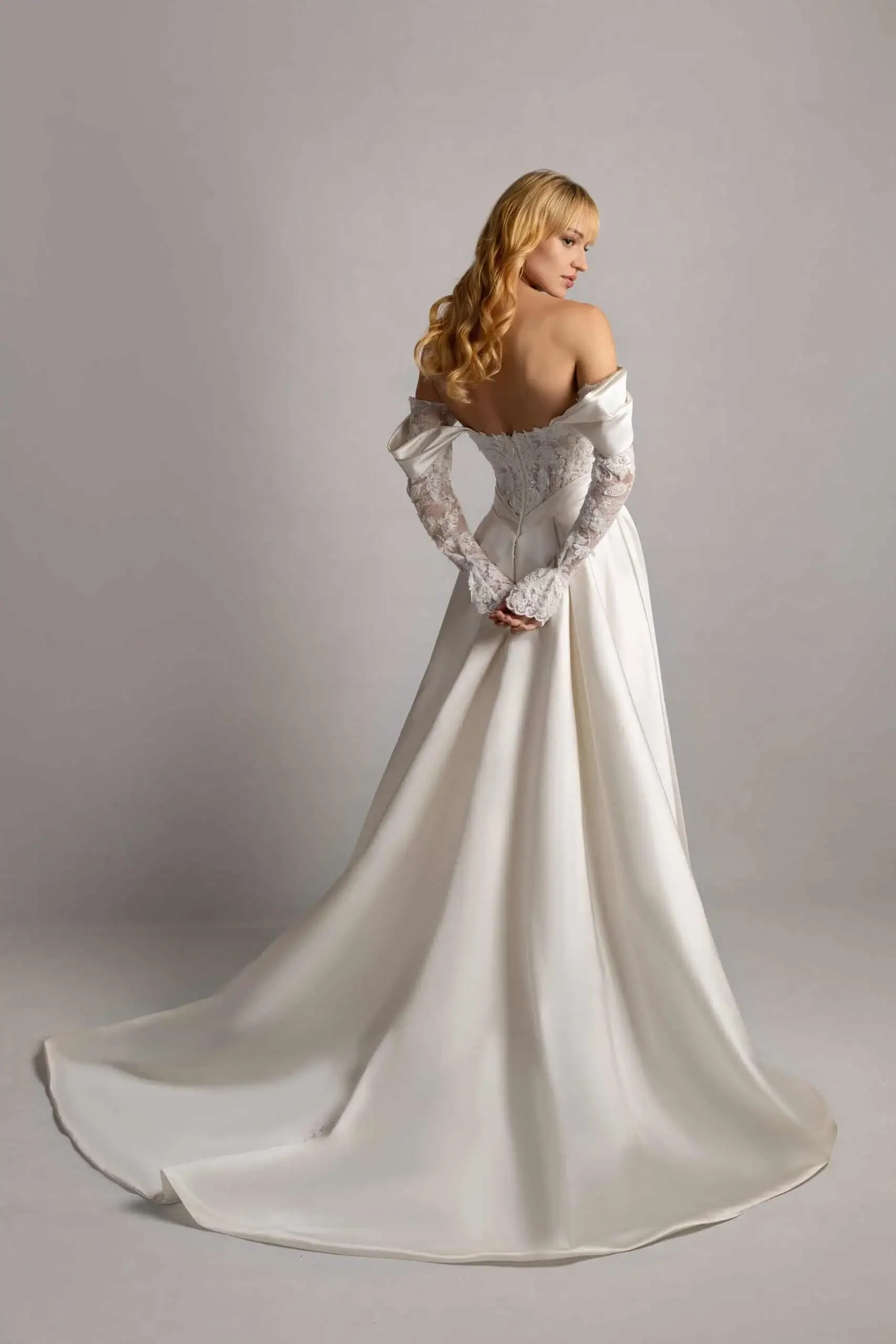 A woman with blonde hair in a wedding dress with lace sleeves and off-the-shoulder design, standing on a plain gray background.