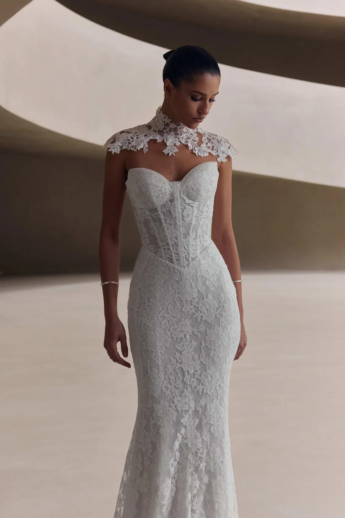 A woman is wearing a fitted white lace wedding dress with a high neck lace overlay and a strapless corset bodice, standing in an abstract modern architectural interior.