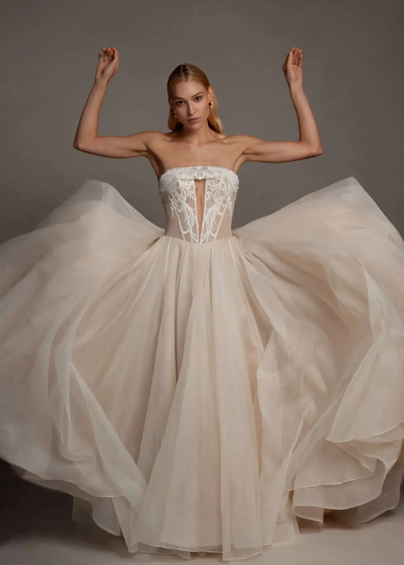 A woman in a strapless, cream-colored wedding gown with intricate lace details on the bodice, standing against a plain background with her arms raised.