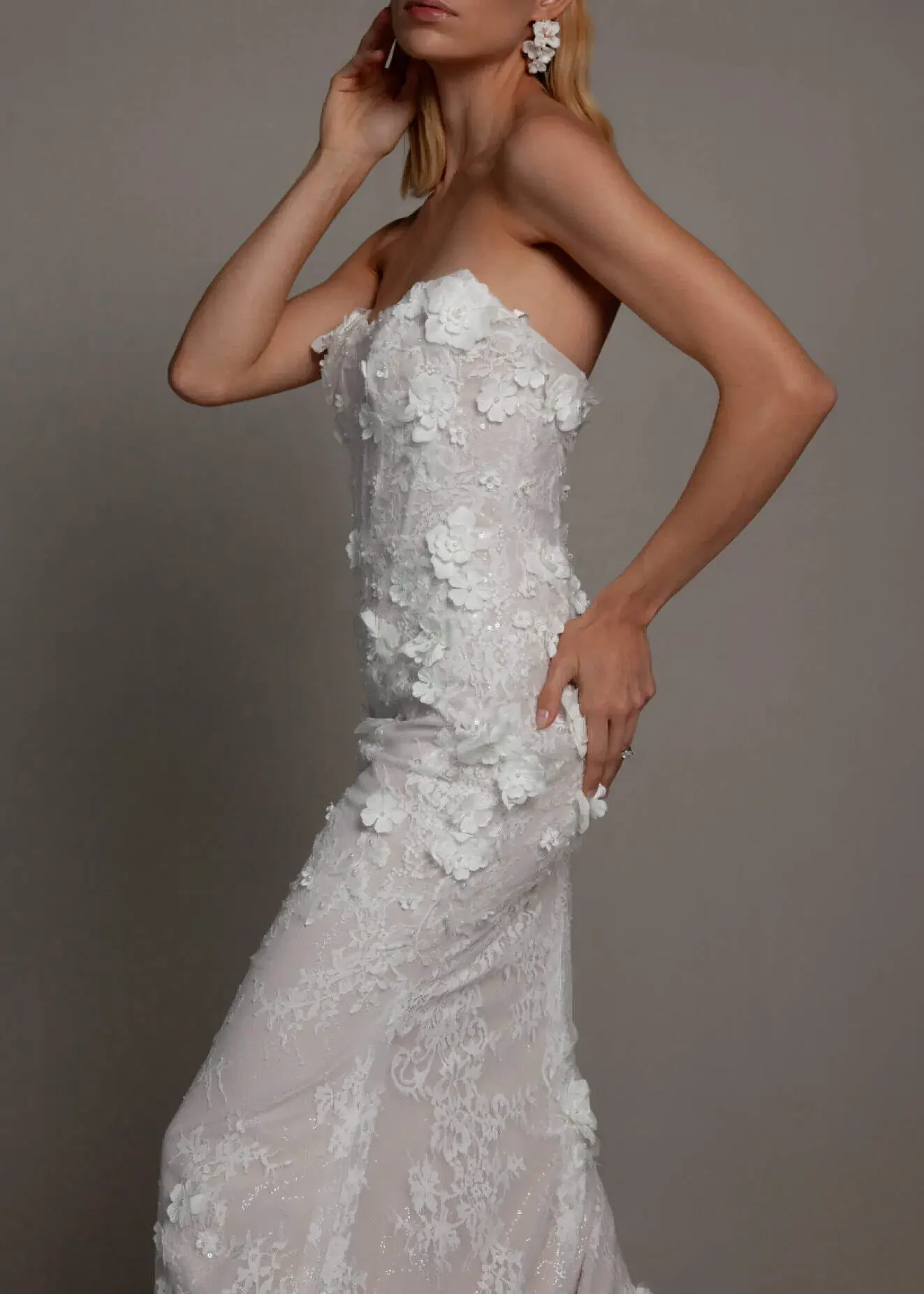 A woman wearing a strapless white lace wedding gown with 3D floral appliqués, standing against a plain gray background.