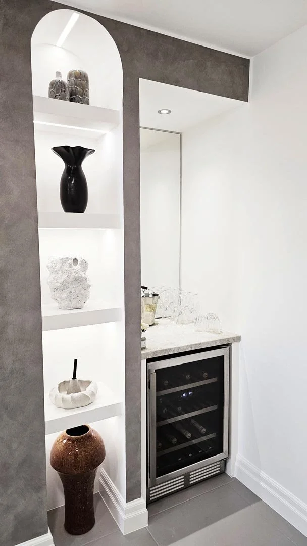 Built-in white shelves with decorative vases and objects, gray wall, wine fridge, countertop with glasses and a bucket, and gray tiled floor.