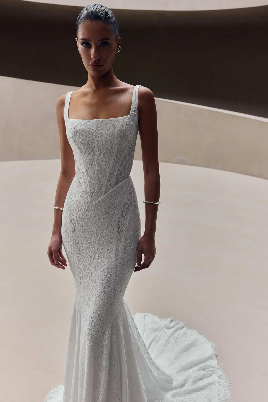 A woman with dark hair styled in a sleek bun wearing a fitted white lace wedding gown with a train.