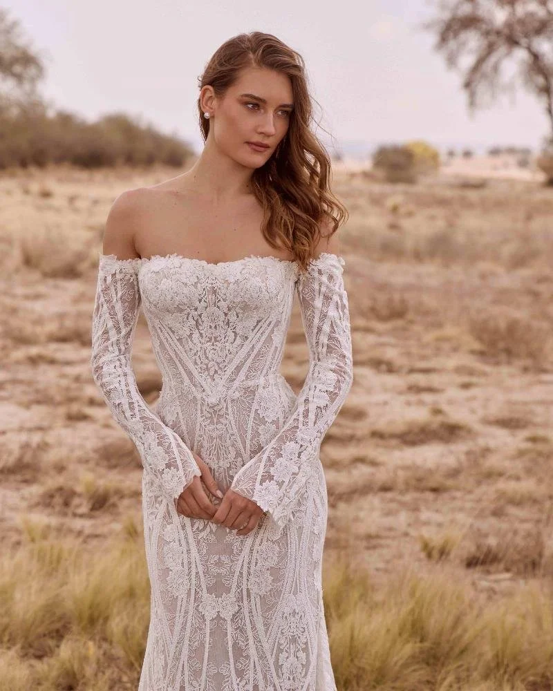 A woman wearing an off-shoulder white lace wedding dress outdoors in a desert landscape.