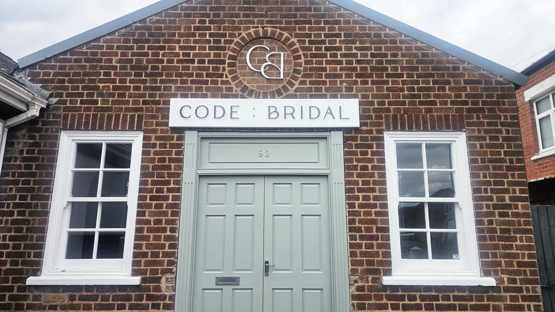Brick building with two white-framed windows, a light gray door, and a sign that reads 'CODE : BRIDAL' above the door.