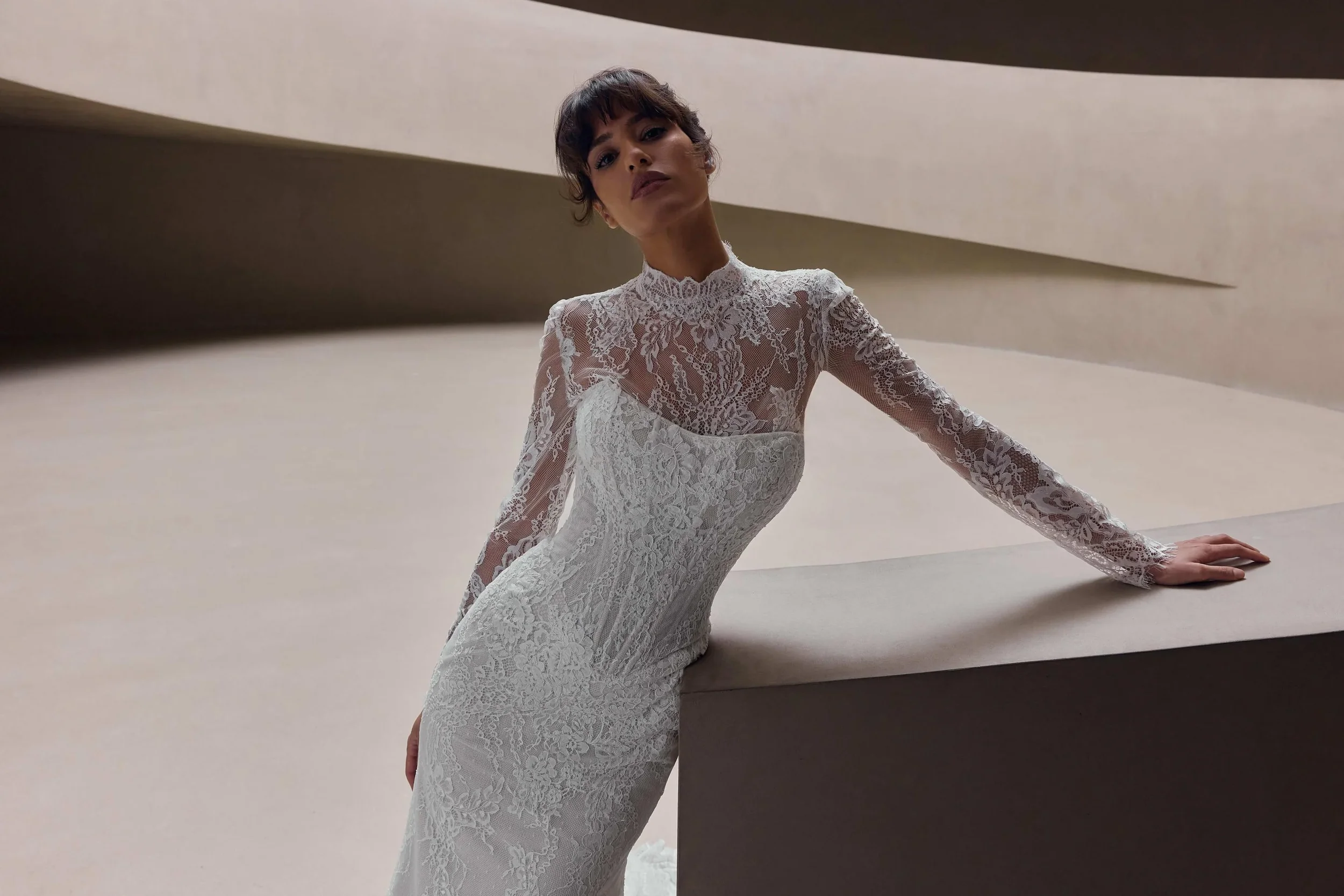 A woman in a white lace dress leaning on a beige curved surface in a modern interior setting.