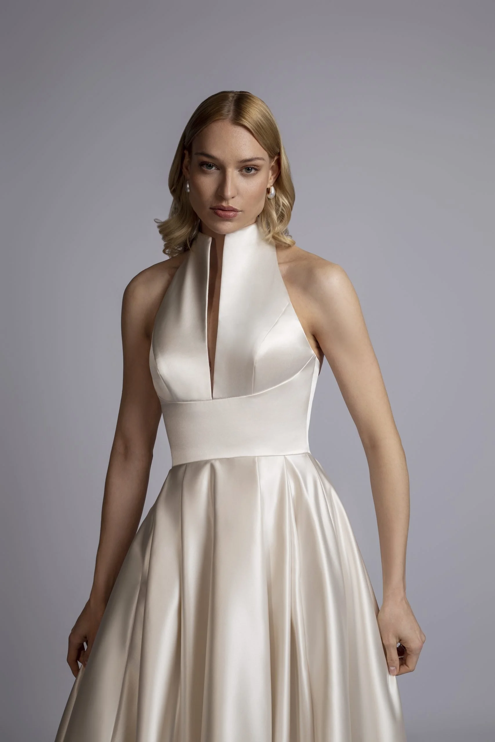 A woman with blonde hair wearing a white satin dress with a halter neck and a fitted waist, standing against a plain gray background.