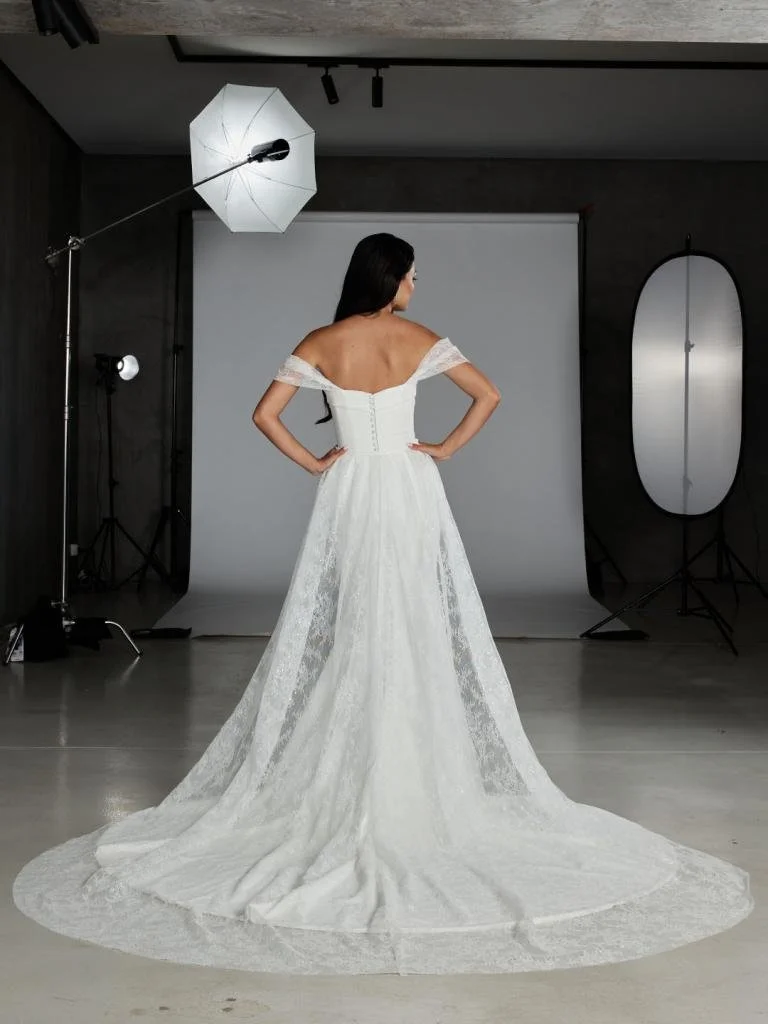 A woman in a white wedding dress standing in a photography studio with her back to the camera.