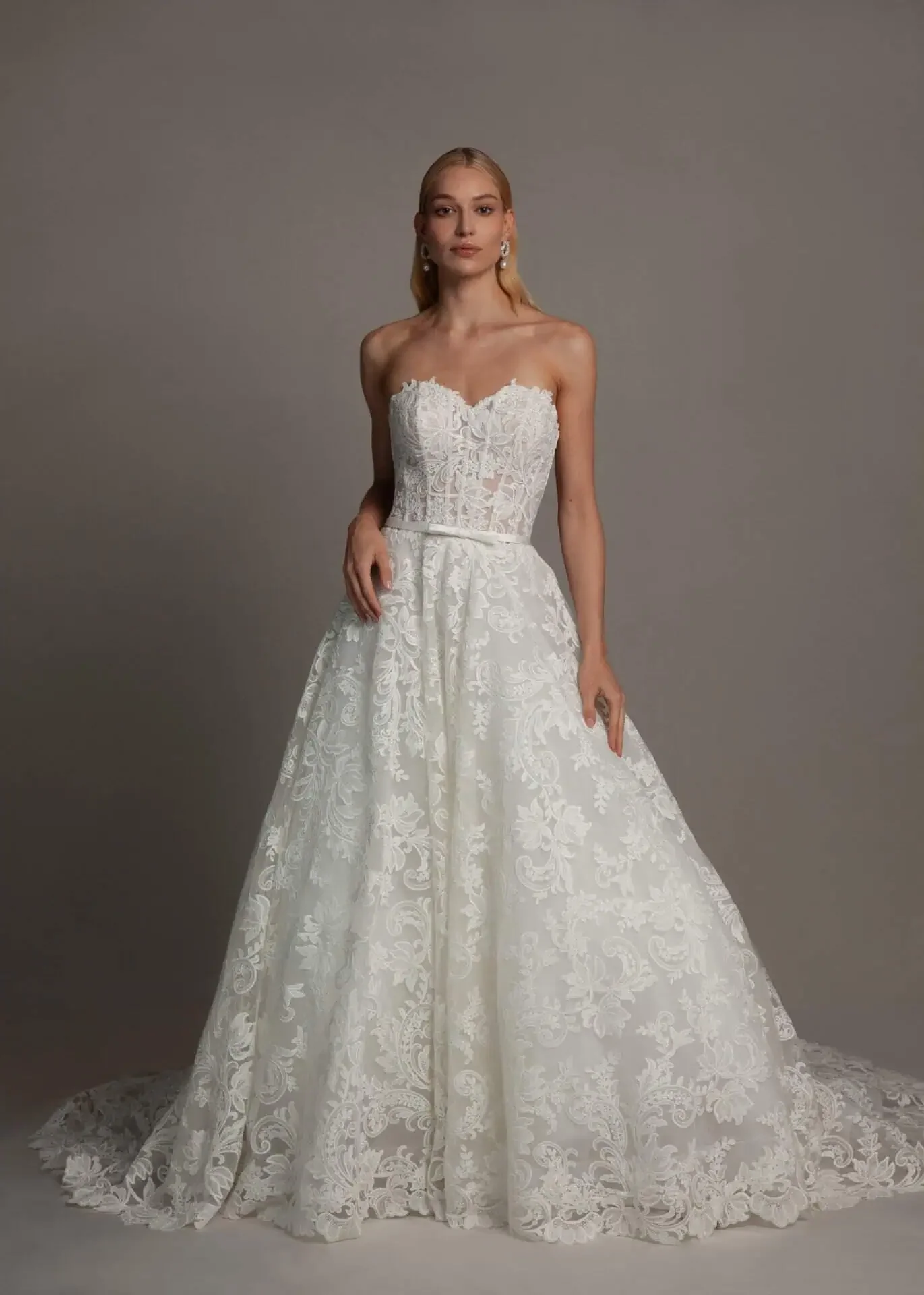 A woman in a strapless white wedding gown with lace detailing, standing against a plain gray background.