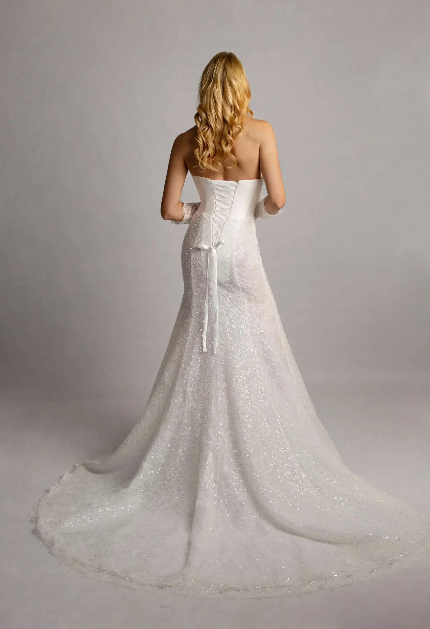 Back view of a woman in a strapless, white wedding gown with a fitted bodice, lace-up back, and a long, glittery train, standing against a plain light gray background.