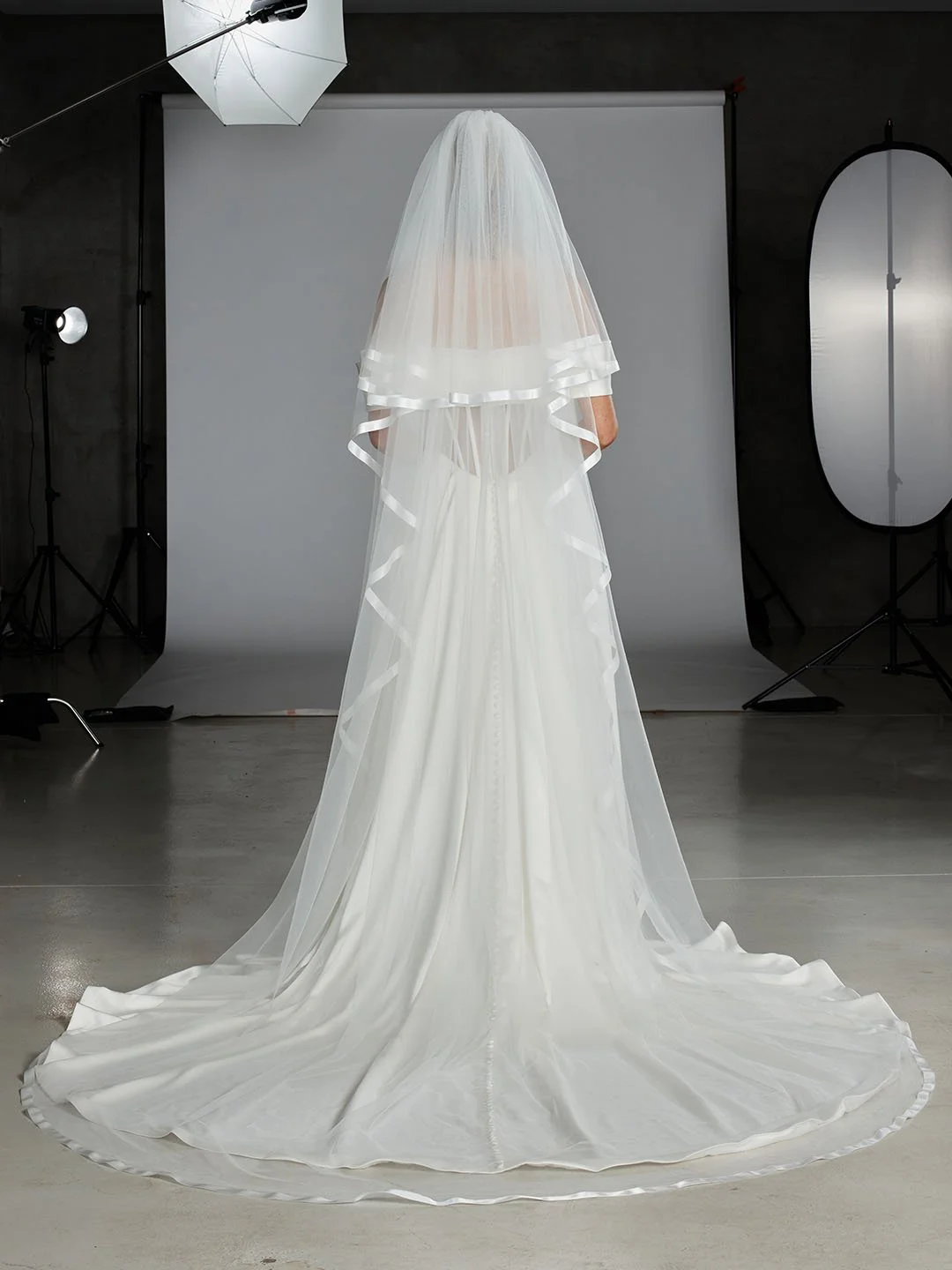 A wedding dress with a long train and a veil, displayed on a mannequin in a photography studio with professional lighting and backdrop.