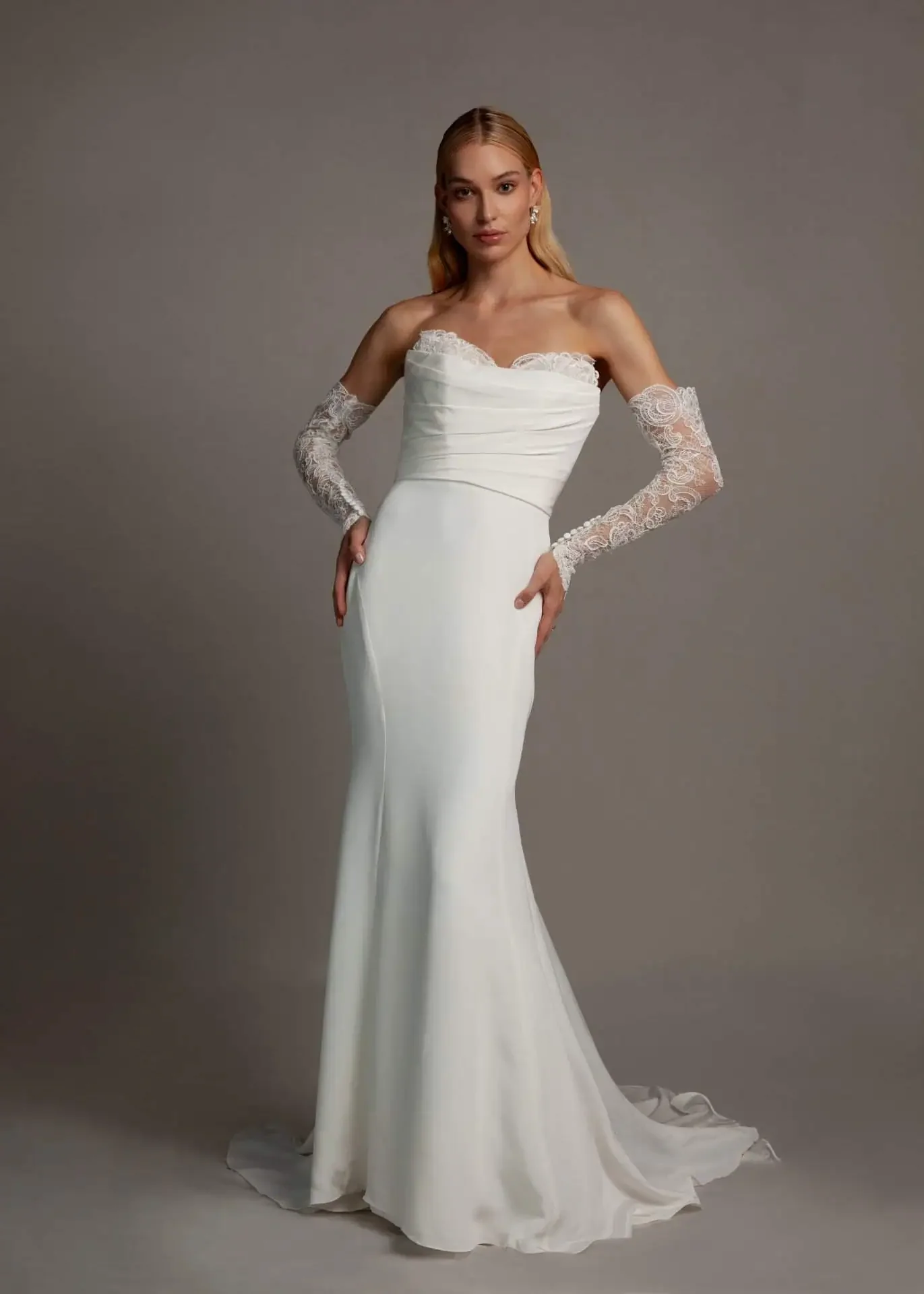 A woman in a white strapless wedding gown with lace gloves standing against a plain gray background.