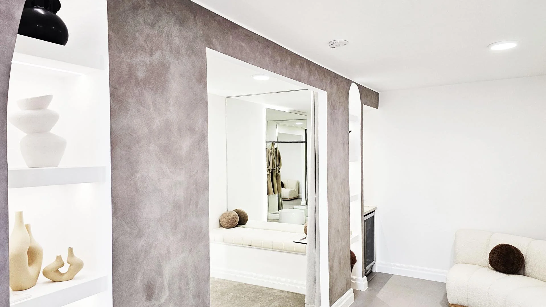 Interior of a modern, minimalist living space with white walls, a white couch with a round brown pillow, and built-in shelves displaying white and beige decorative vases.