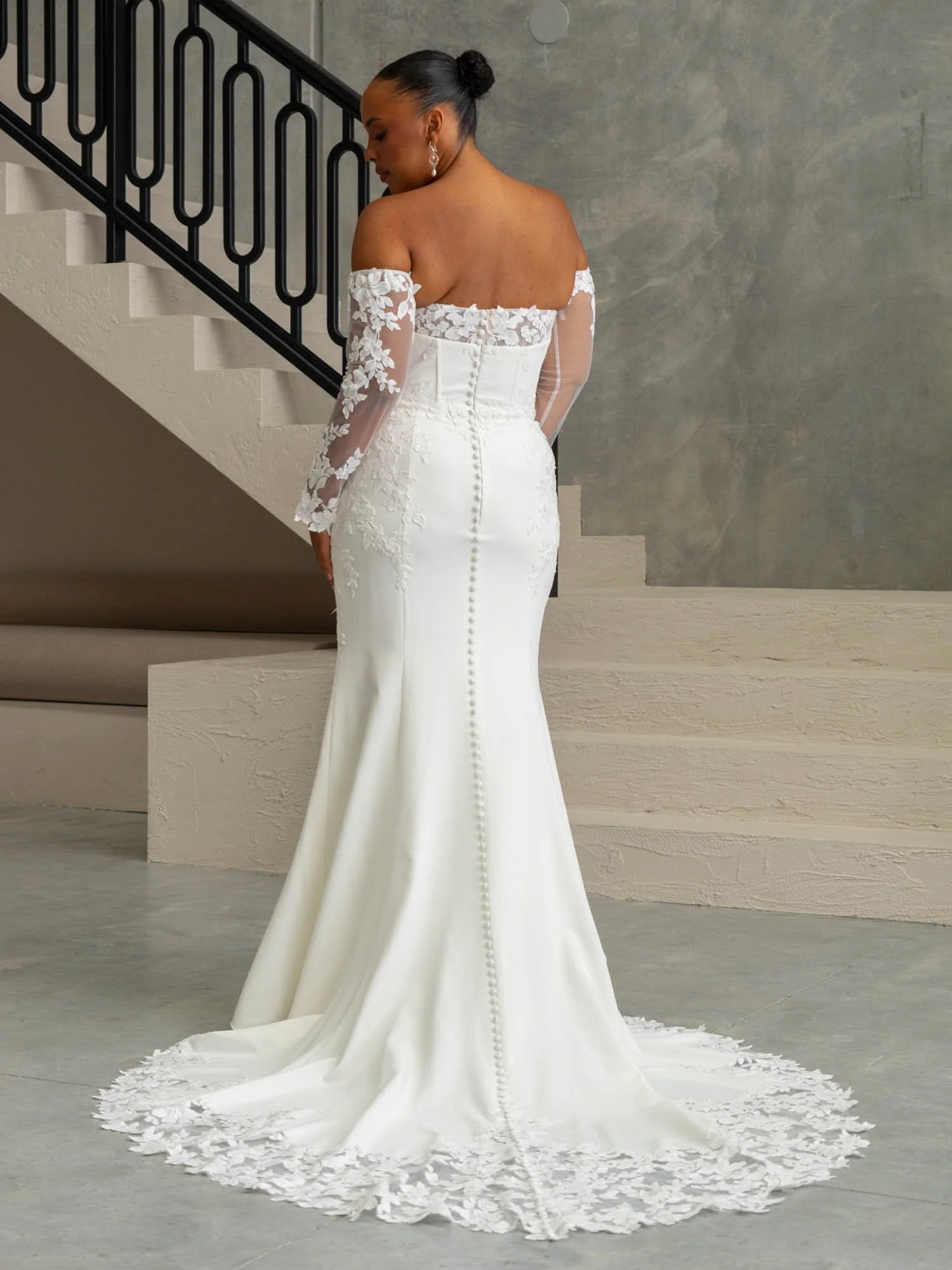 A woman in a white wedding gown with lace off-the-shoulder sleeves, standing near a staircase with black railings, facing away from the camera.