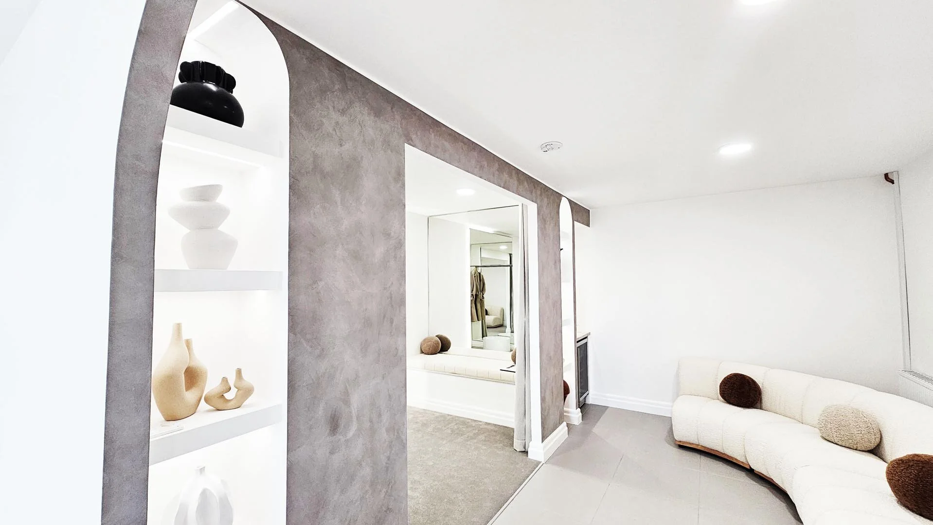 Modern interior living space with minimalist decor, white curved sofa with round pillows, open shelving with vases and bowls, neutral color palette, and a seating nook with a carpet.