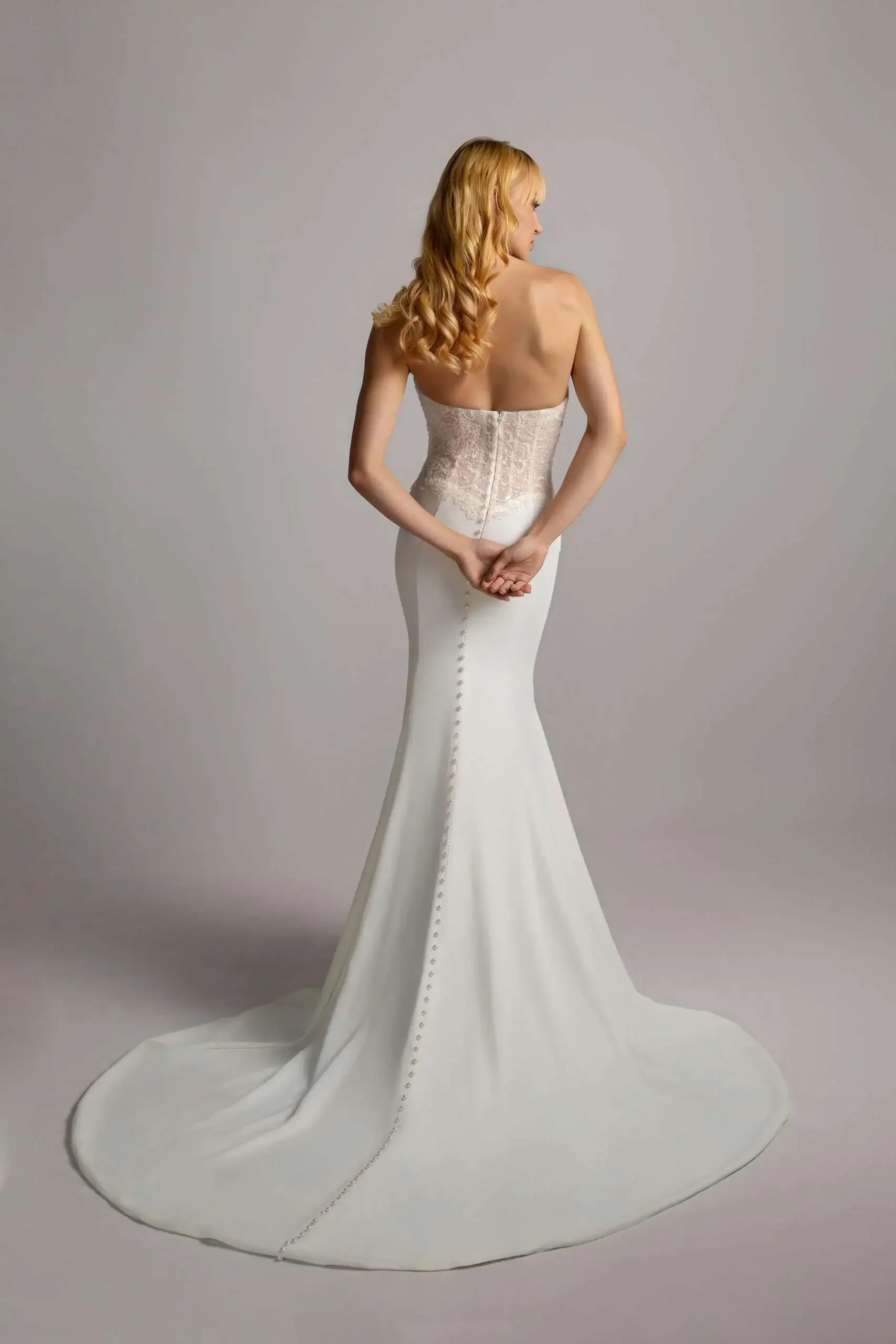 A woman with long, wavy red hair is wearing a strapless white bridal gown with a fitted bodice and a long train, standing with her back to the camera against a plain gray background.