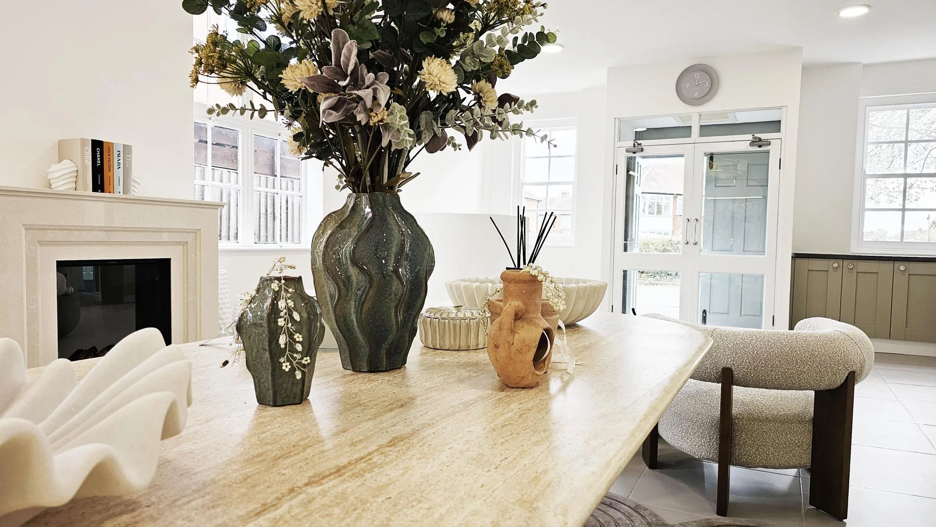 A dining room with a wooden table decorated with vases and bowls, beige chairs, large windows providing natural light, and a white fireplace with books on top.