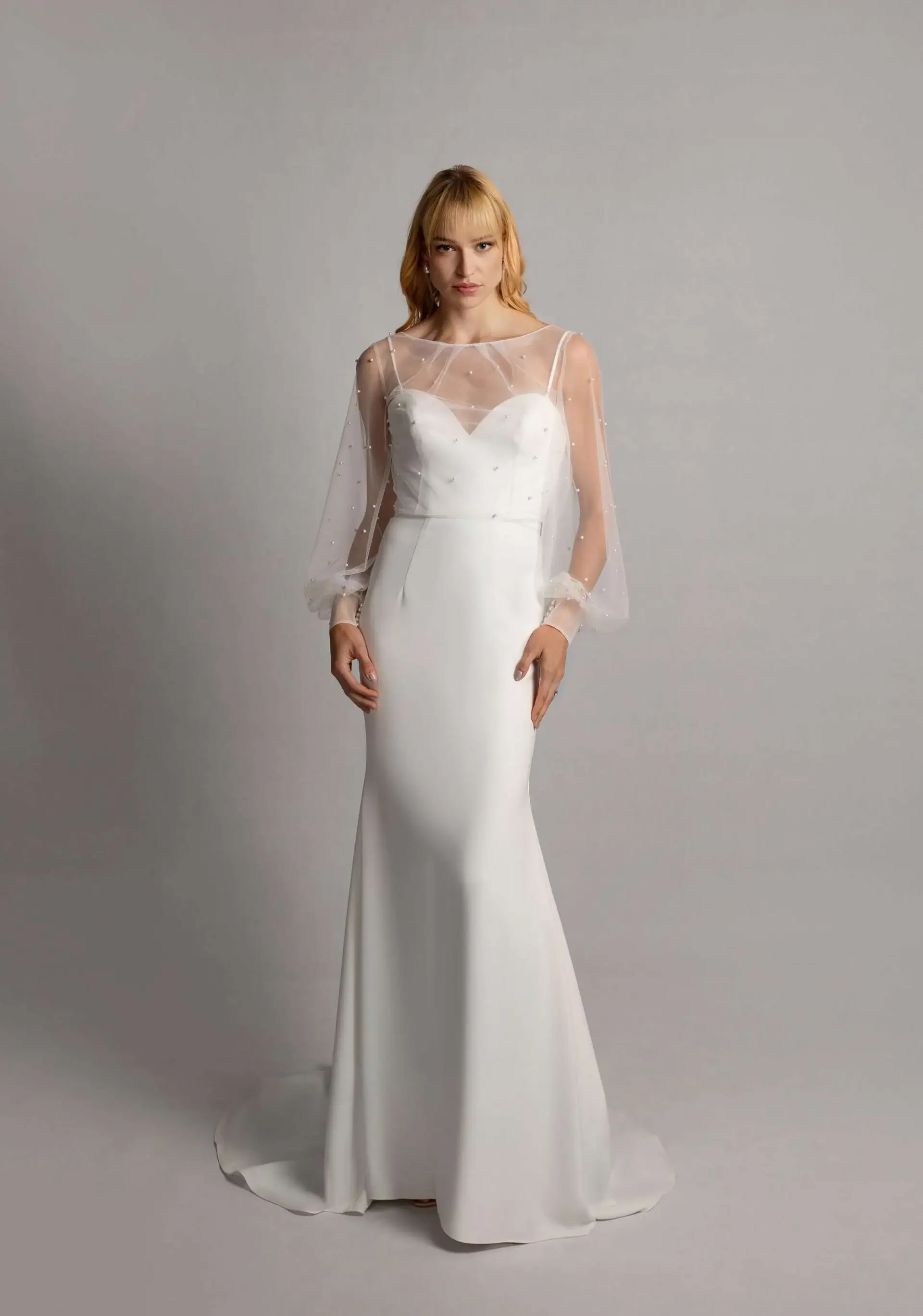 A woman in a white wedding gown with a fitted silhouette, wearing a sheer, beaded cape over her shoulders, stands against a plain gray background.