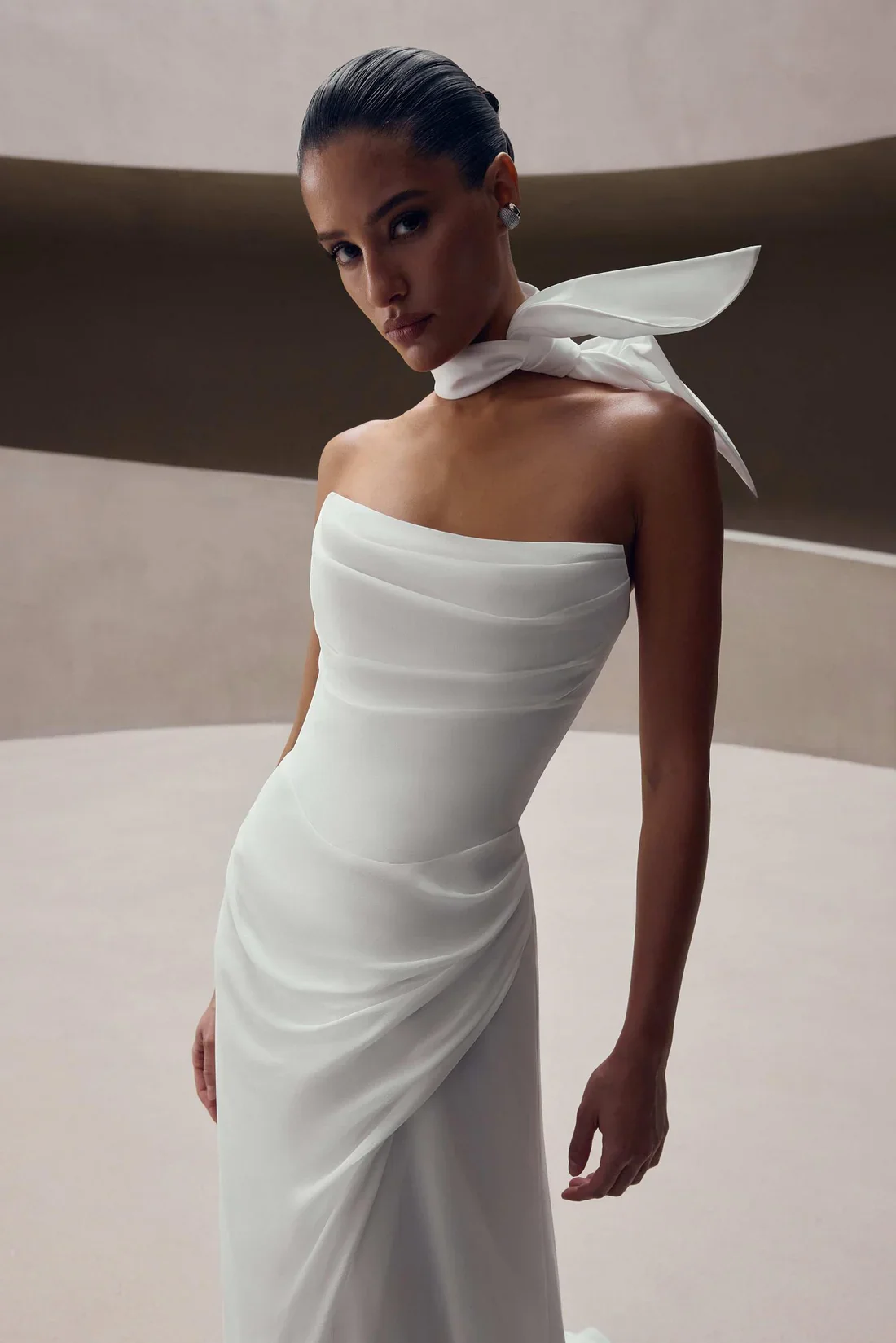A woman in a strapless white dress with a draped bodice and a high slit, accessorized with earrings and a white scarf tied around her neck, standing against a modern, minimalist background.