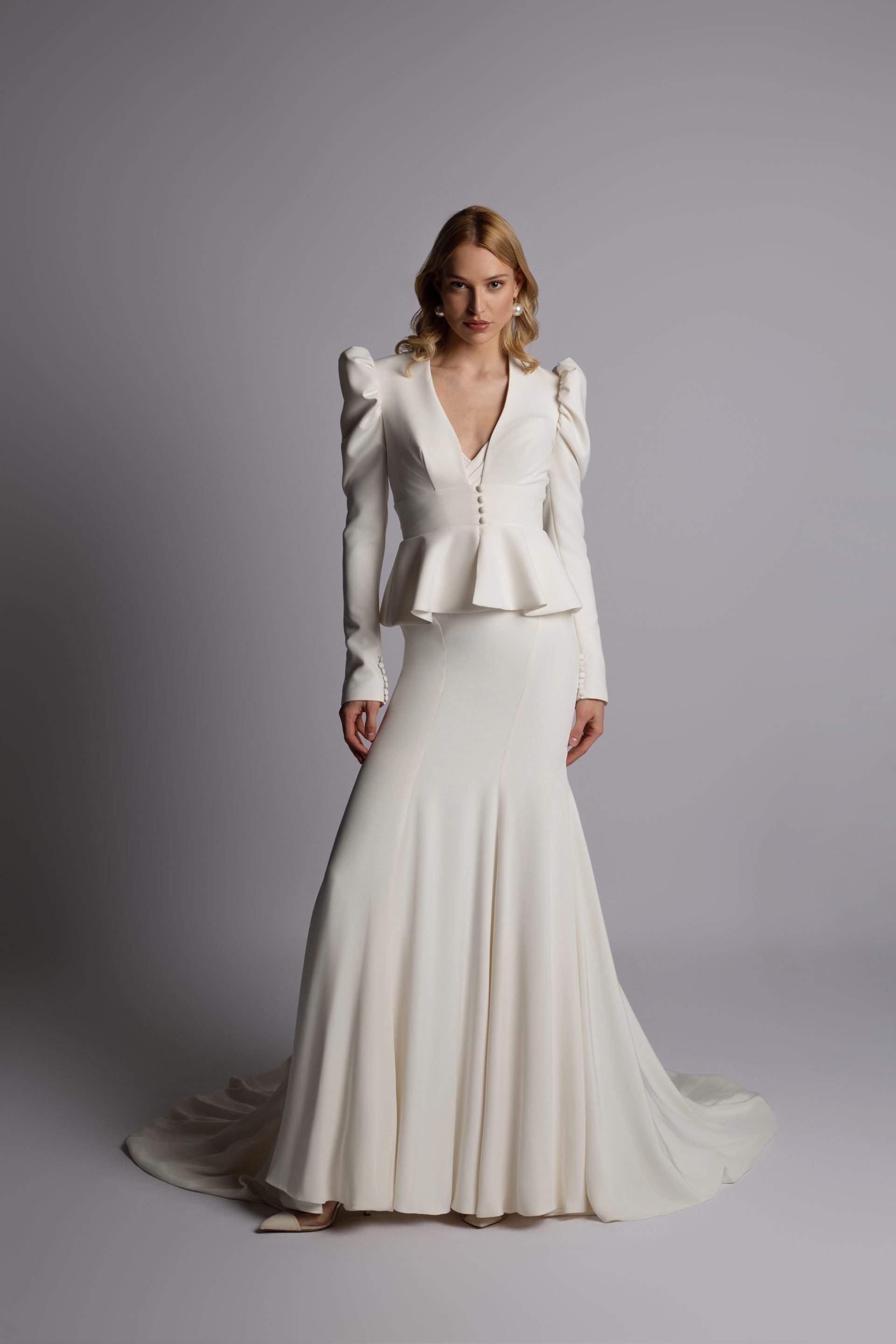 A woman in an elegant white gown with puffed sleeves and a fitted waist, standing on a plain background.