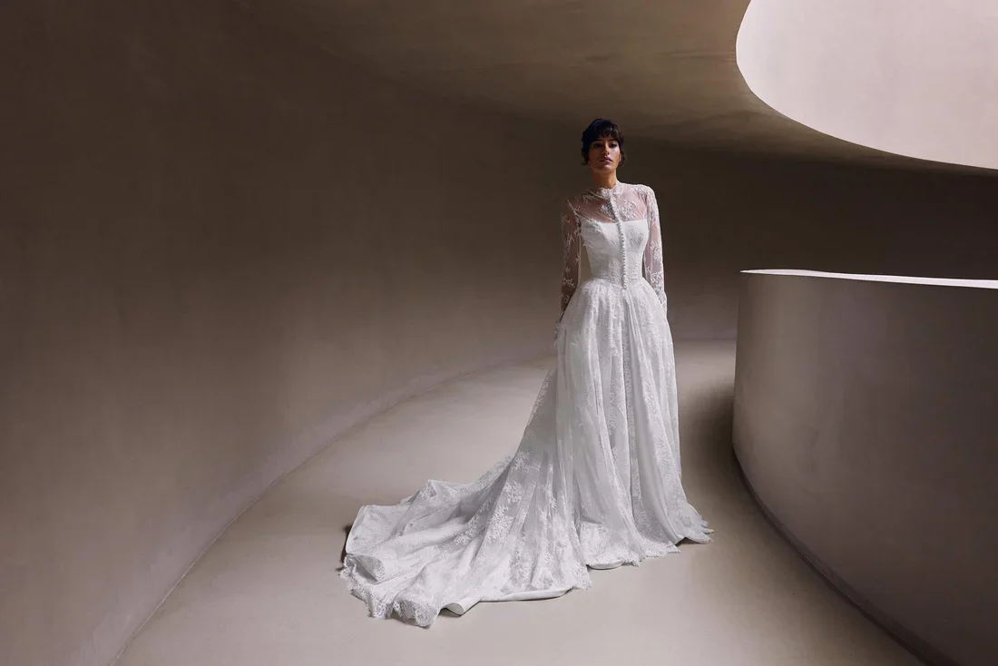 A woman in a long white lace wedding dress with long sleeves, standing in a minimalistic, modern, curved concrete space.