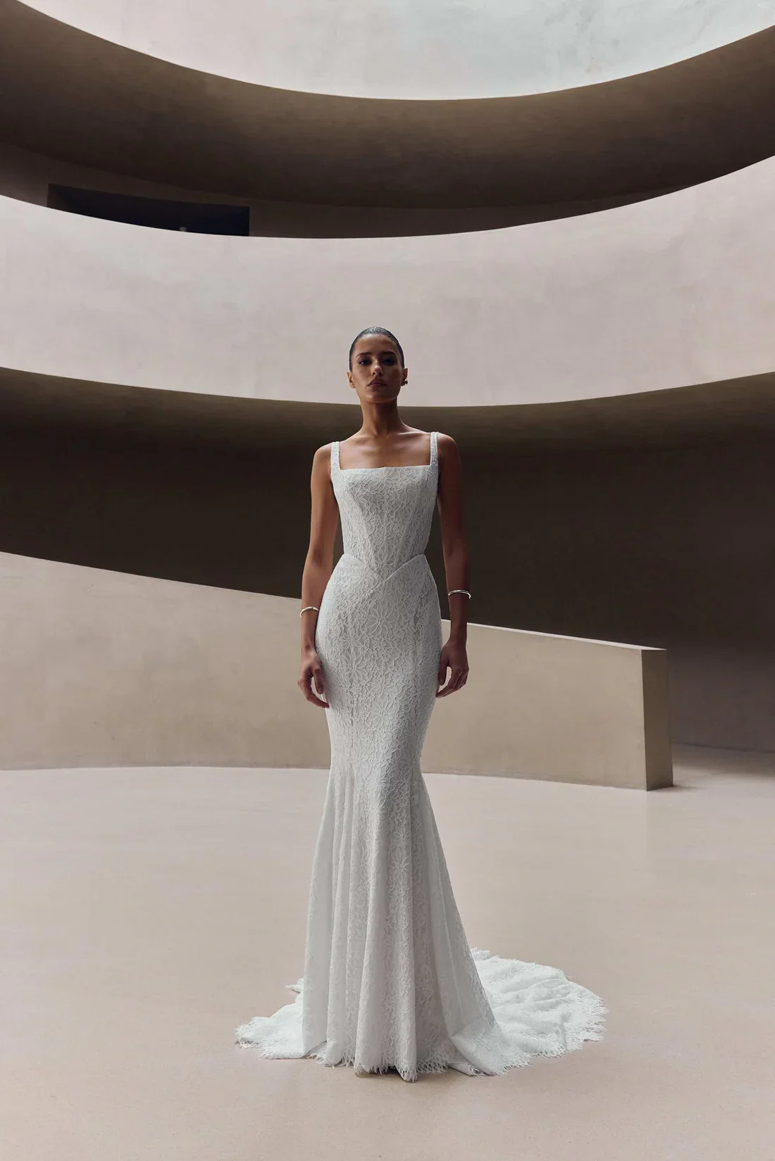 A woman in a white lace dress standing in a modern, minimalist architectural space with curved beige walls and ceiling openings.
