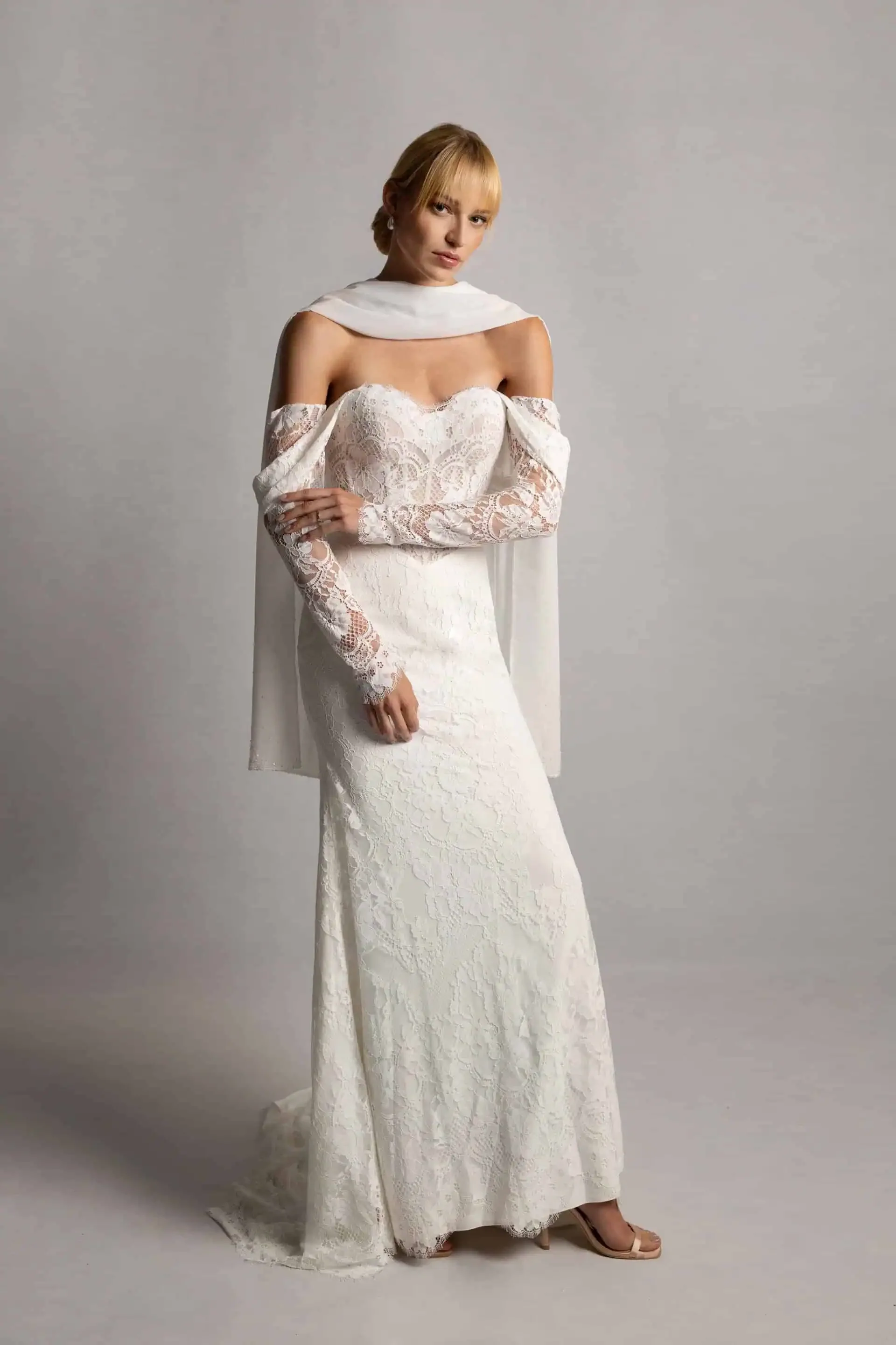 A woman in a white lace wedding gown with off-the-shoulder sleeves and matching lace gloves, standing against a plain gray background.