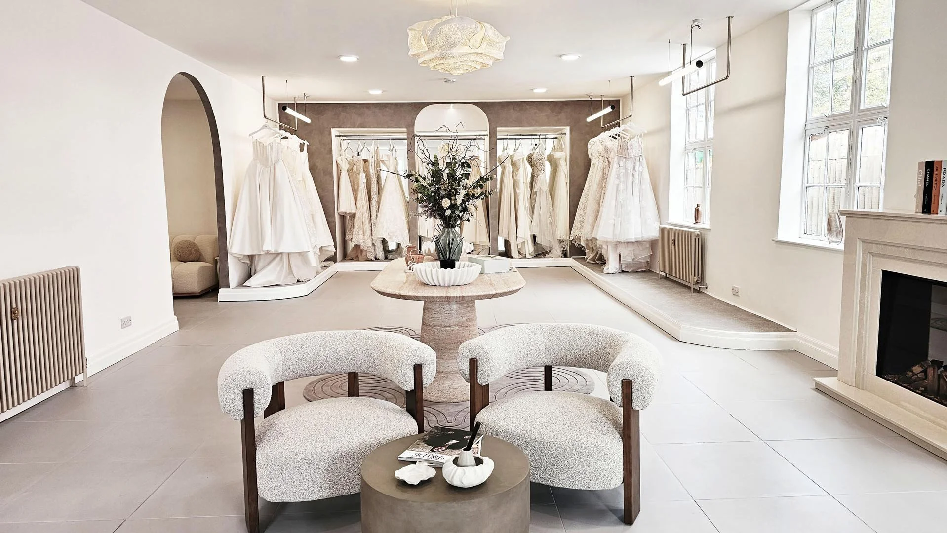 Interior of a bridal boutique with a display of wedding dresses on racks, a central table with a flower arrangement, upholstered seating, and a fireplace.