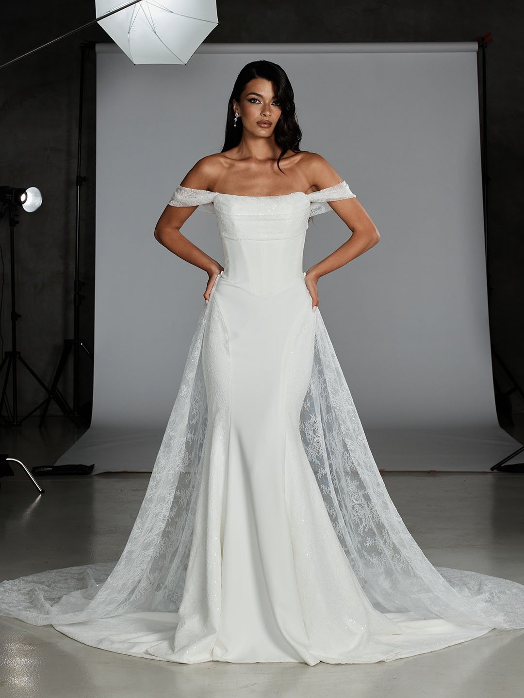 Woman modeling a white off-the-shoulder wedding dress with lace details and a train, in a photography studio.