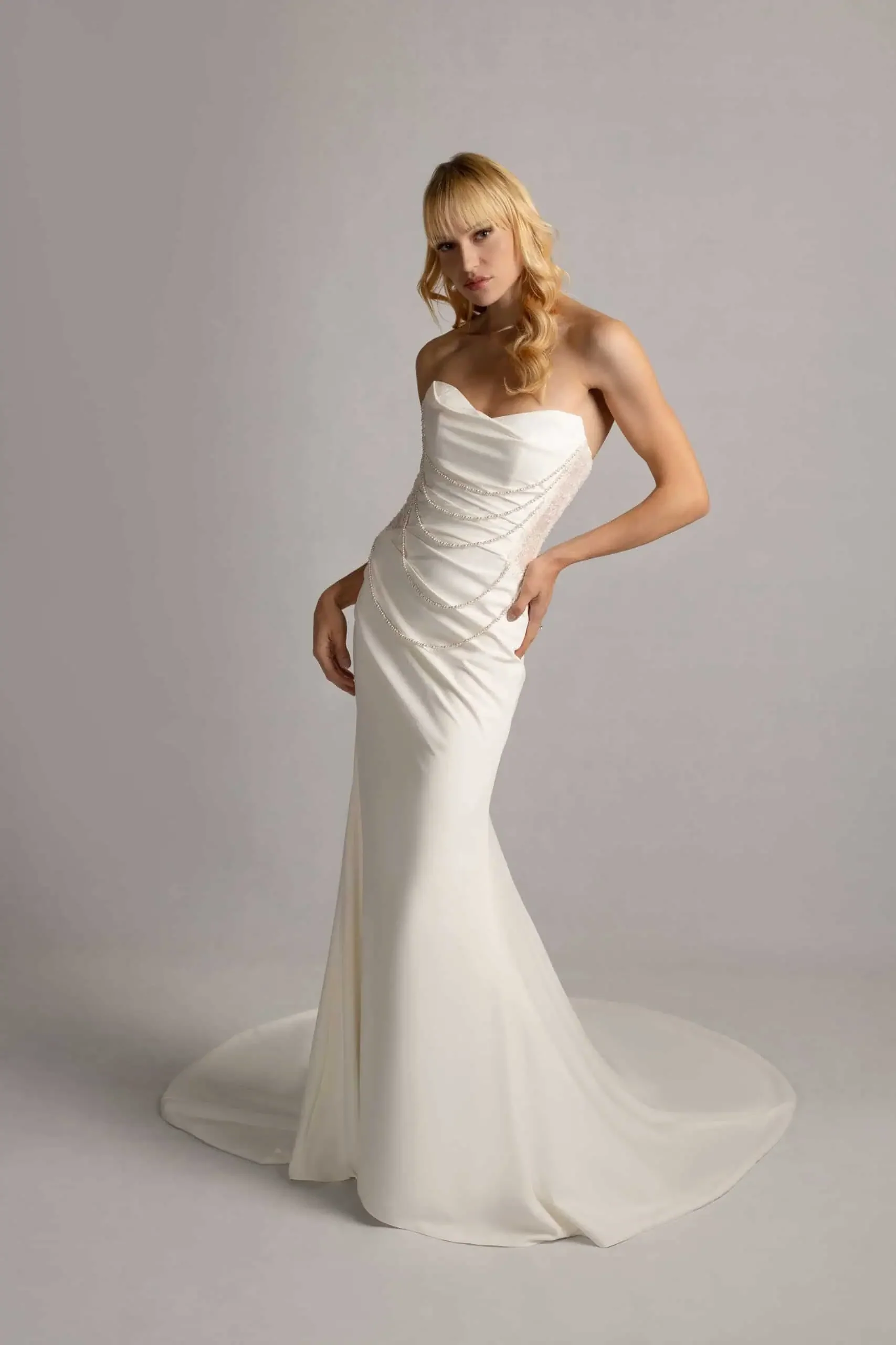 A woman in a strapless white satin wedding dress with pearl embellishments, standing against a plain gray background.