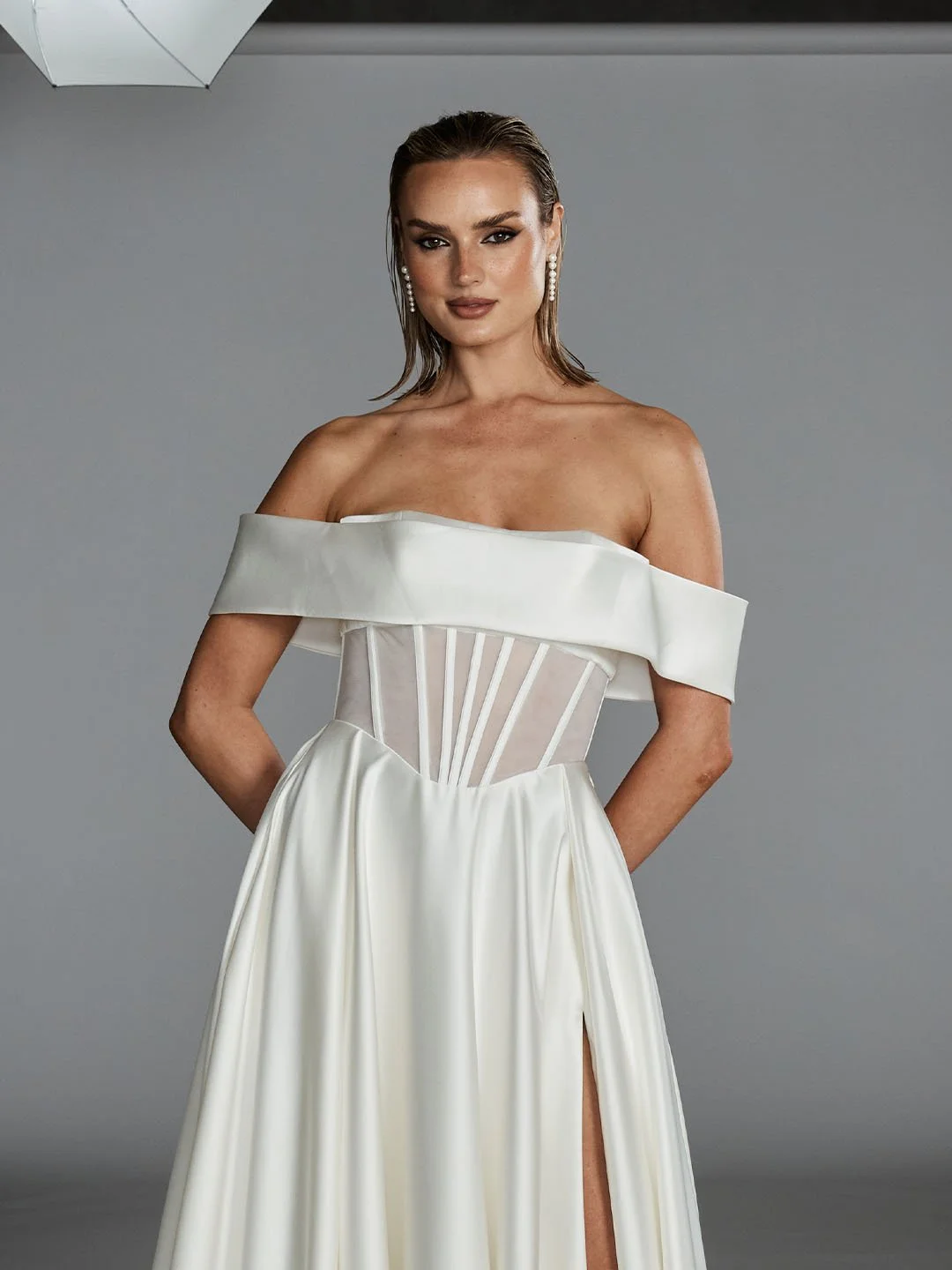 Woman in elegant off-shoulder white dress with sheer corset detail, standing against a gray background, wearing pearl earrings.