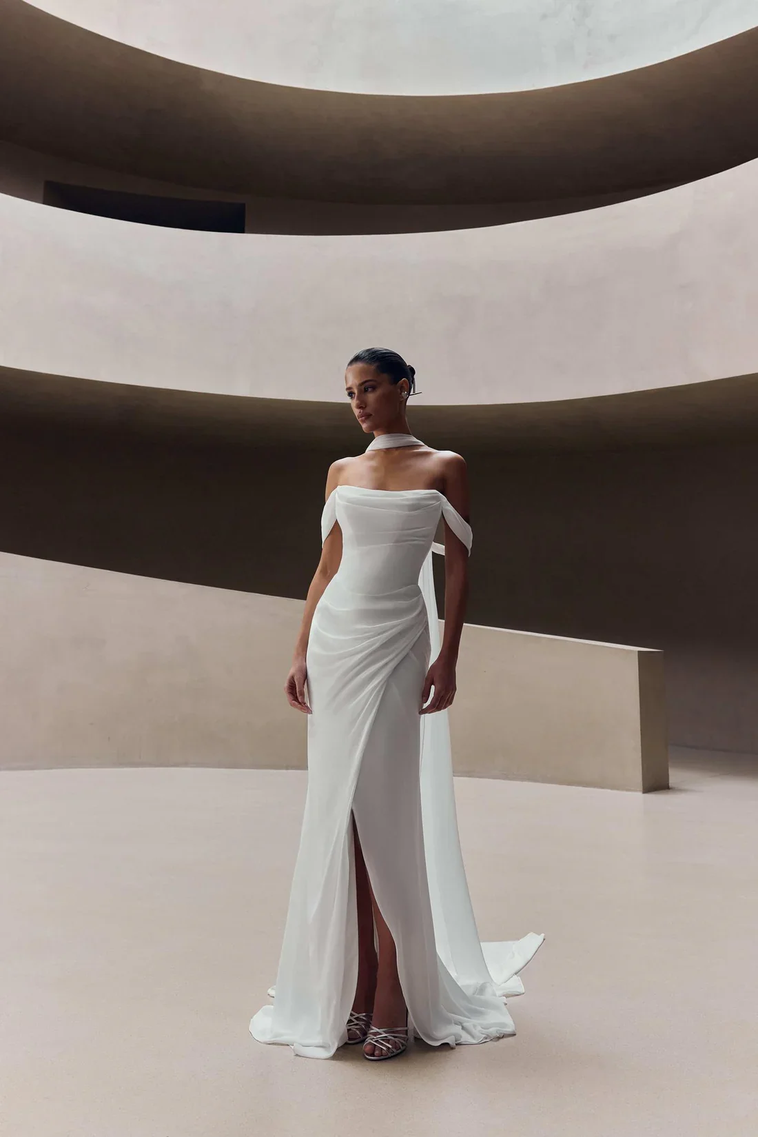 A woman in a white gown standing in a minimalistic, modern architectural space with curved walls and natural light.